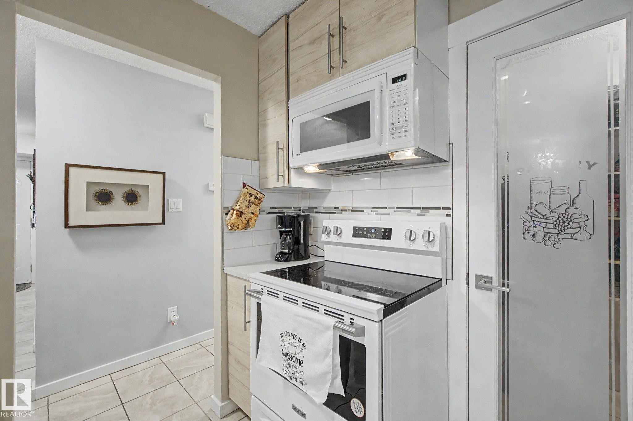 Kitchen featuring white appliances, a white tile backsplash, light wood-finish cabinetry, a built-in microwave, and an adjacent frosted glass pantry door - #39 3111 142 Av Nw, Edmonton, AB - Indoor Photo Showing Kitchen