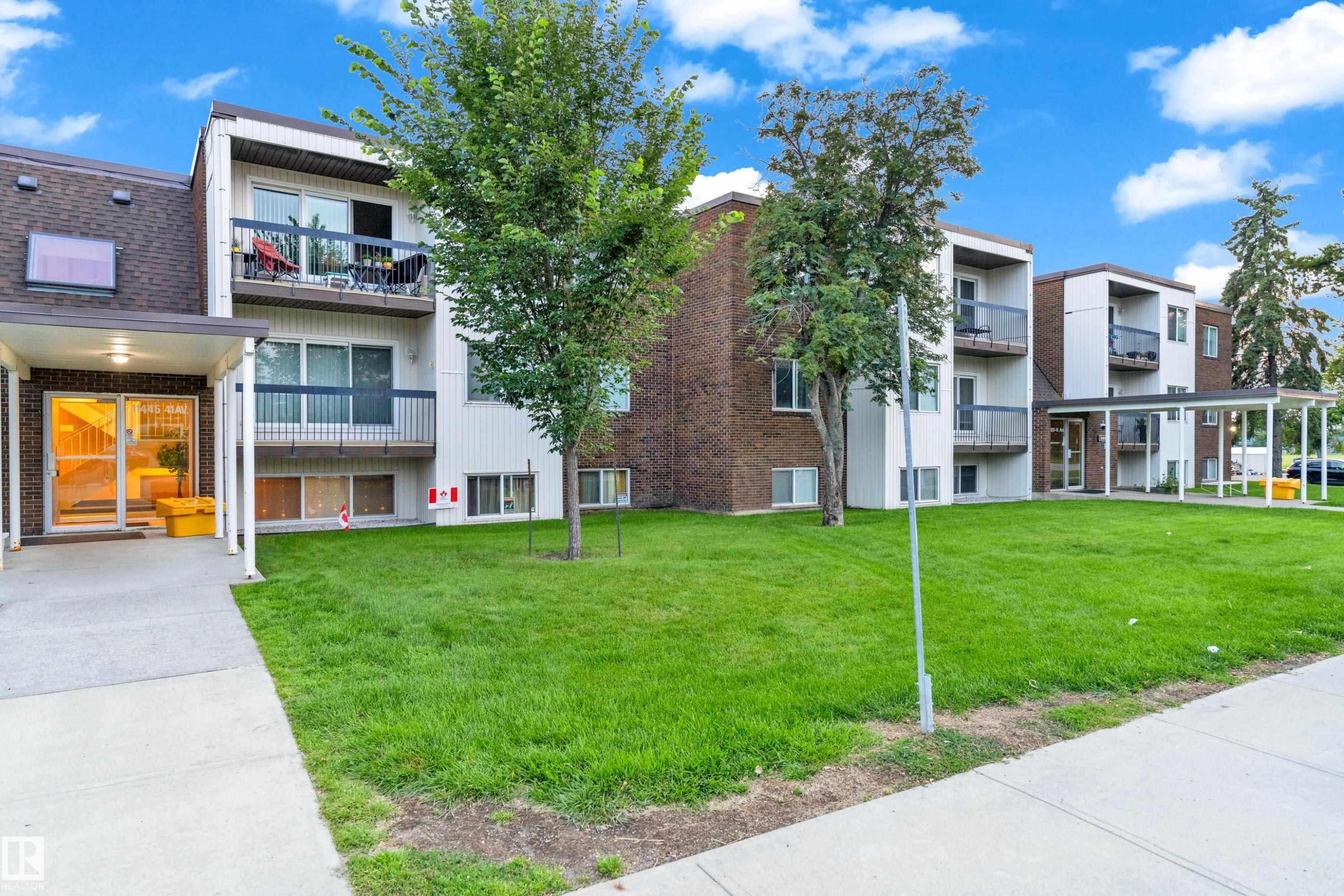 Multi-story building featuring brick and white siding, individual balconies with dark railings, a covered entrance, and a landscaped lawn - #104 11445 41 Av Nw, Edmonton, AB - Outdoor