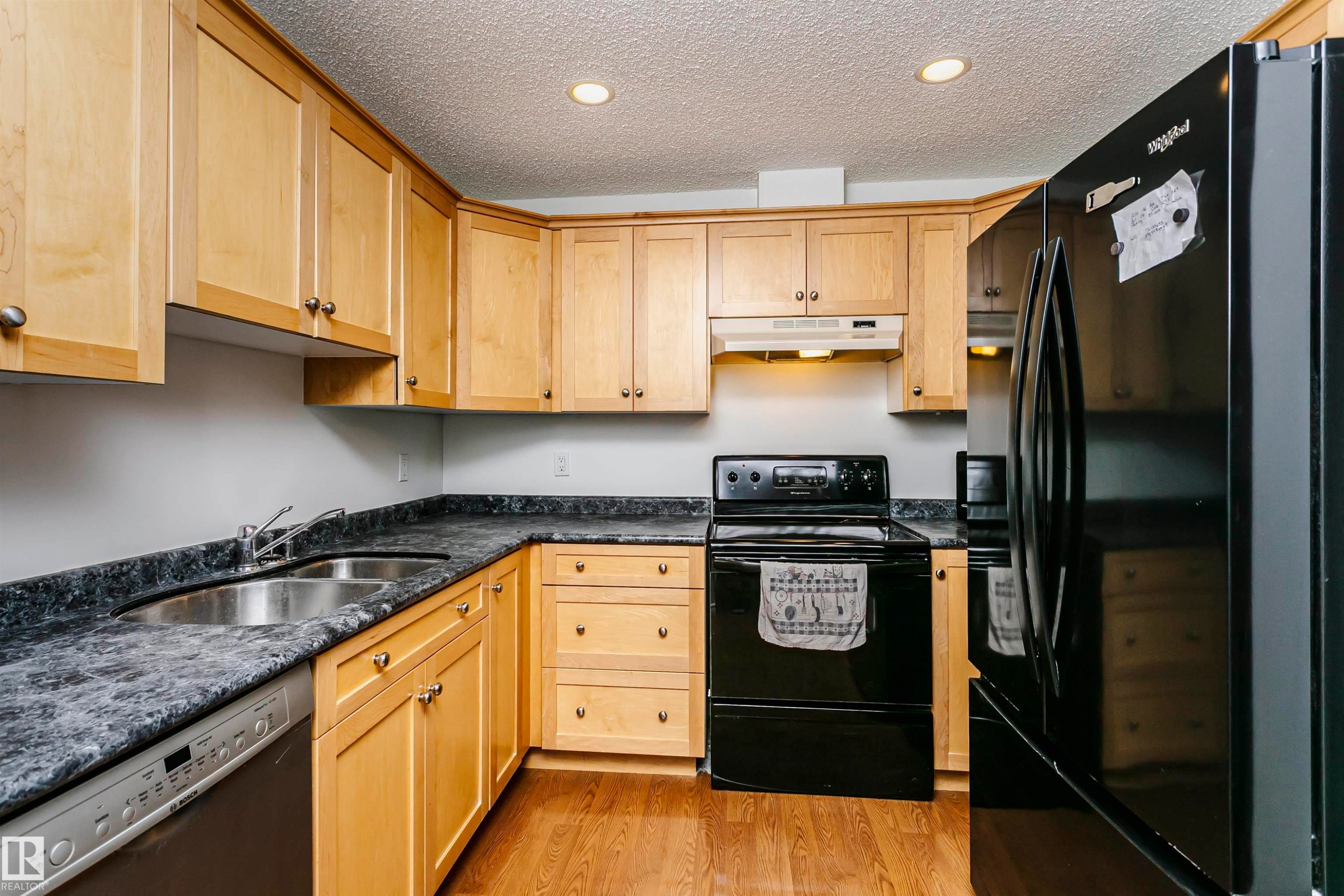 Kitchen featuring light wood cabinetry with brushed nickel hardware, black appliances, a double basin stainless steel sink, and wood-finish flooring - #106 12110 106 Av Nw, Edmonton, AB - Indoor Photo Showing Kitchen With Double Sink