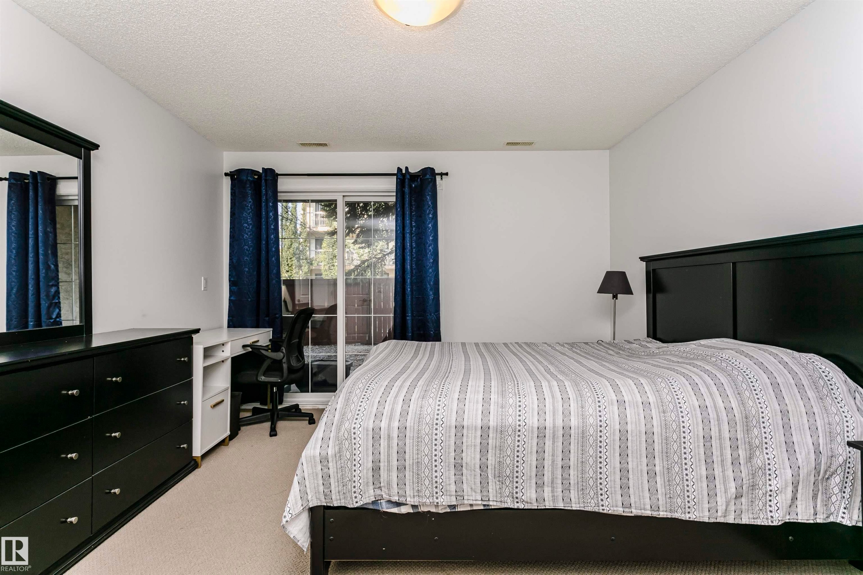 Carpeted room with white walls, featuring a sliding glass door, overhead lighting, and a built-in desk with shelving - #106 12110 106 Av Nw, Edmonton, AB - Indoor Photo Showing Bedroom
