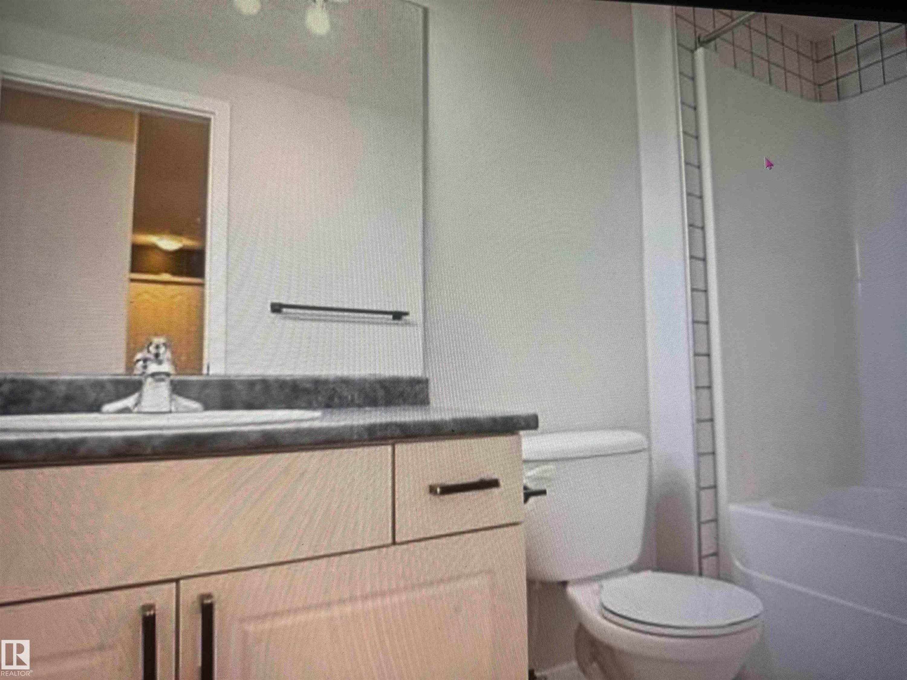 Bathroom featuring a light-toned vanity with dark hardware, a dark countertop, an integrated sink with a chrome faucet, a large wall mirror, and a white toilet - #106 592 Hooke Rd Nw, Edmonton, AB - Indoor Photo Showing Bathroom