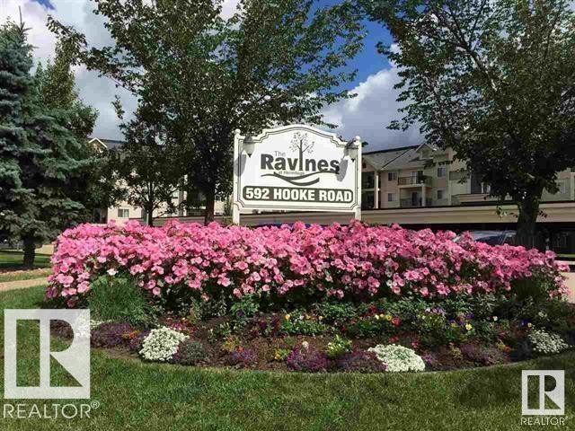 Entry monument with a vibrant floral display featuring pink blooms, evergreen trees, and deciduous trees - #106 592 Hooke Rd Nw, Edmonton, AB - Outdoor
