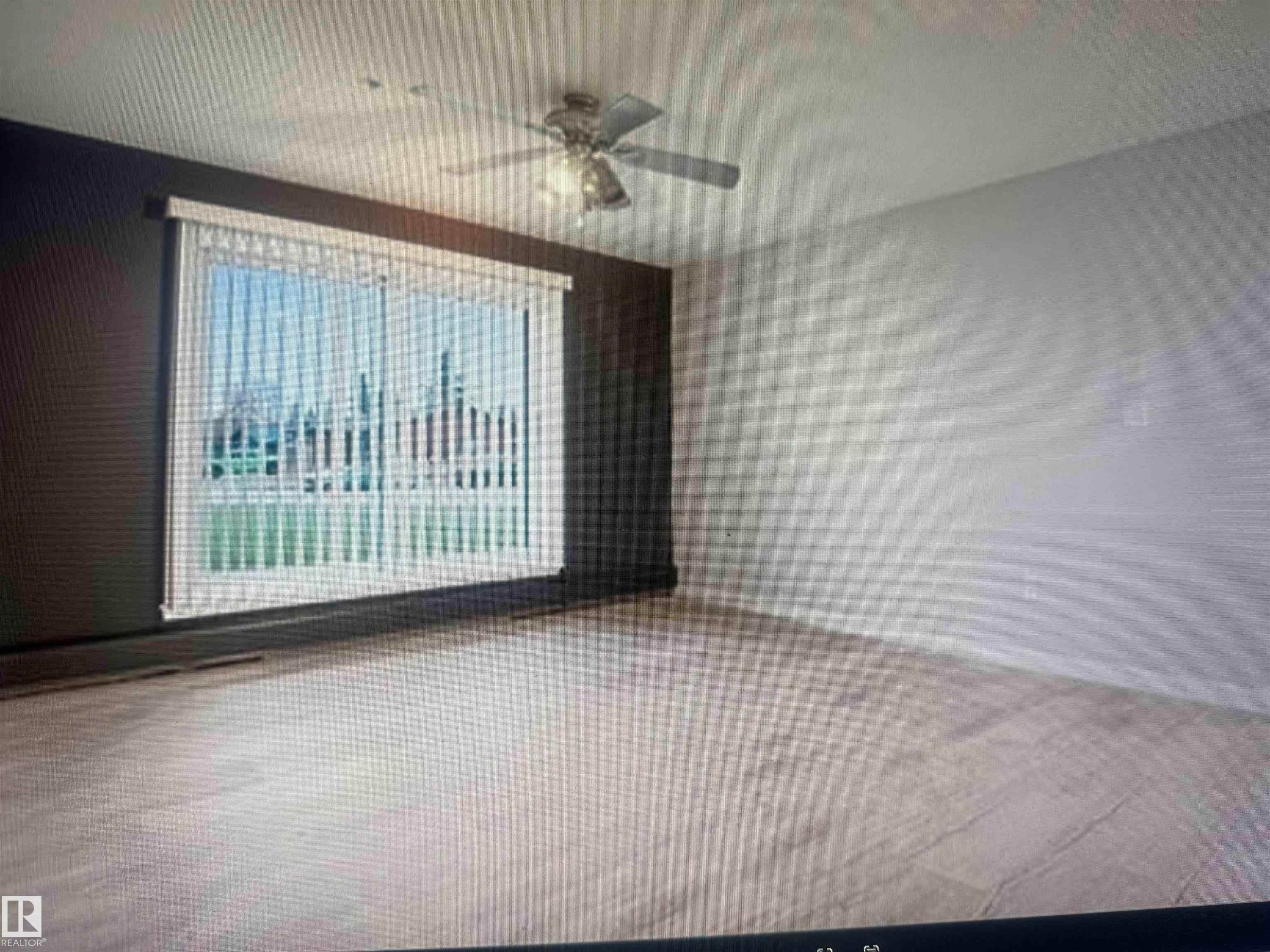 Spacious room with wood-finish flooring, a large window with vertical blinds, and a ceiling fan - #106 592 Hooke Rd Nw, Edmonton, AB - Indoor Photo Showing Other Room