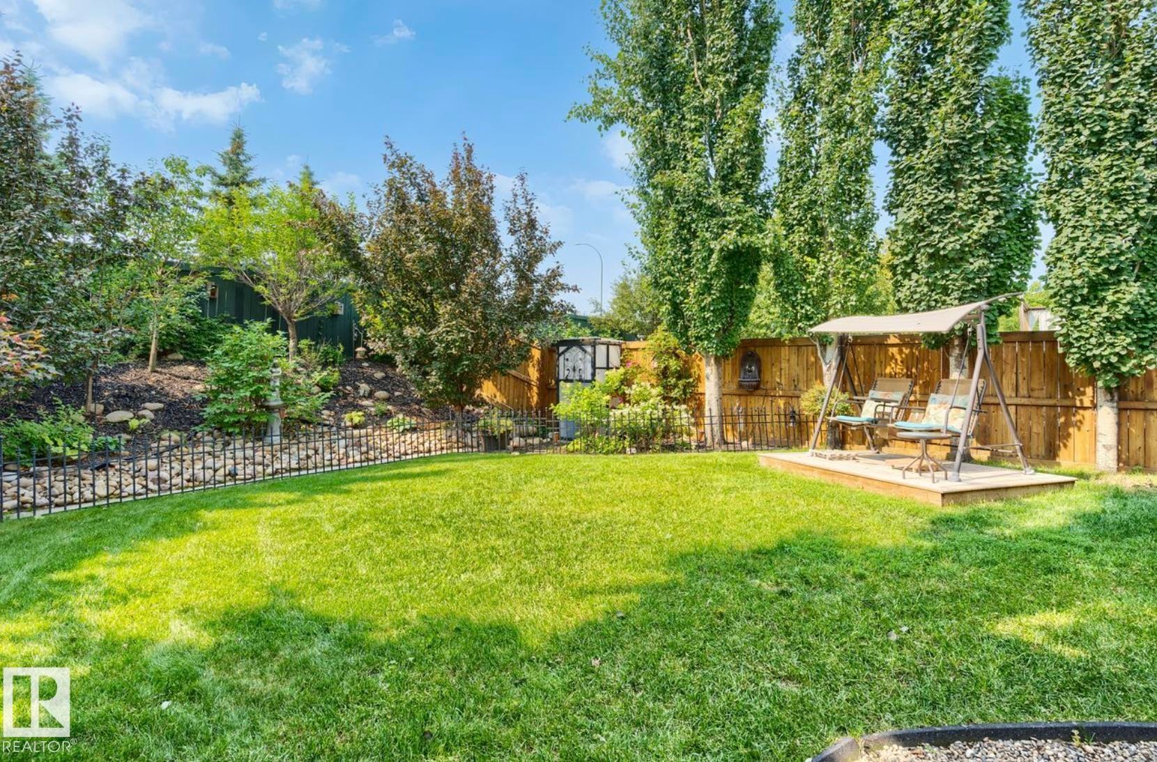Expansive backyard oasis featuring a meticulously manicured lawn, mature landscaping with towering evergreen trees, decorative metal fencing, wooden pergola structure with seating area, and hardscaped pathways - 22 Voleta Crest, Spruce Grove, AB - Outdoor With Backyard
