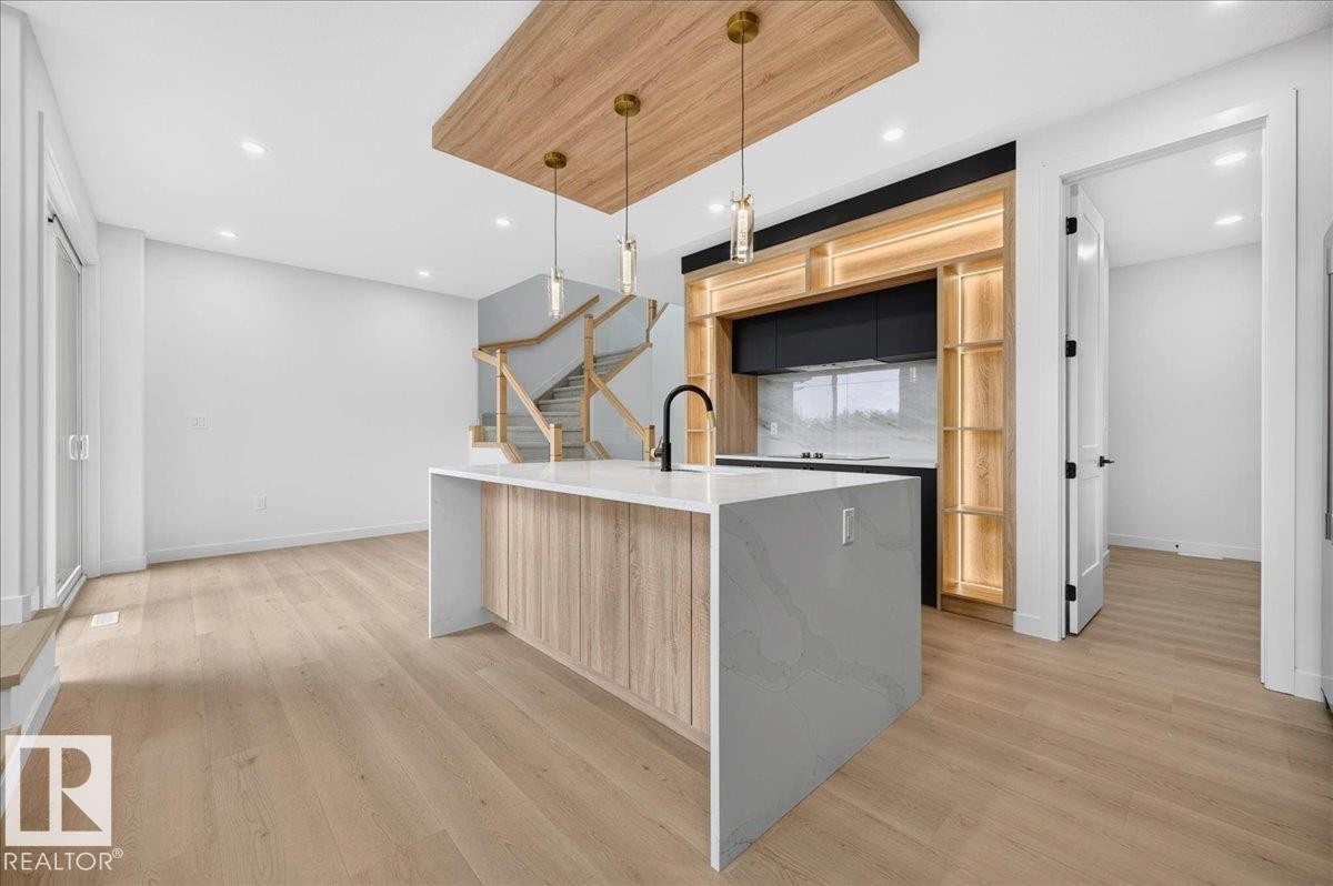 Open-concept living area featuring light wood-finish flooring, a contemporary kitchen island with a waterfall countertop, integrated cabinetry with accent lighting, and a staircase with light wood railings - 403 172 Avenue Nw, Edmonton, AB - Indoor Photo Showing Kitchen