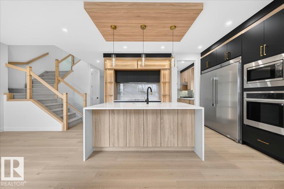 Open-concept kitchen with a large central island, light wood-finish flooring, and a contemporary staircase featuring glass panels and natural wood railings - 403 172 Avenue Nw, Edmonton, AB - Indoor Photo Showing Kitchen With Upgraded Kitchen
