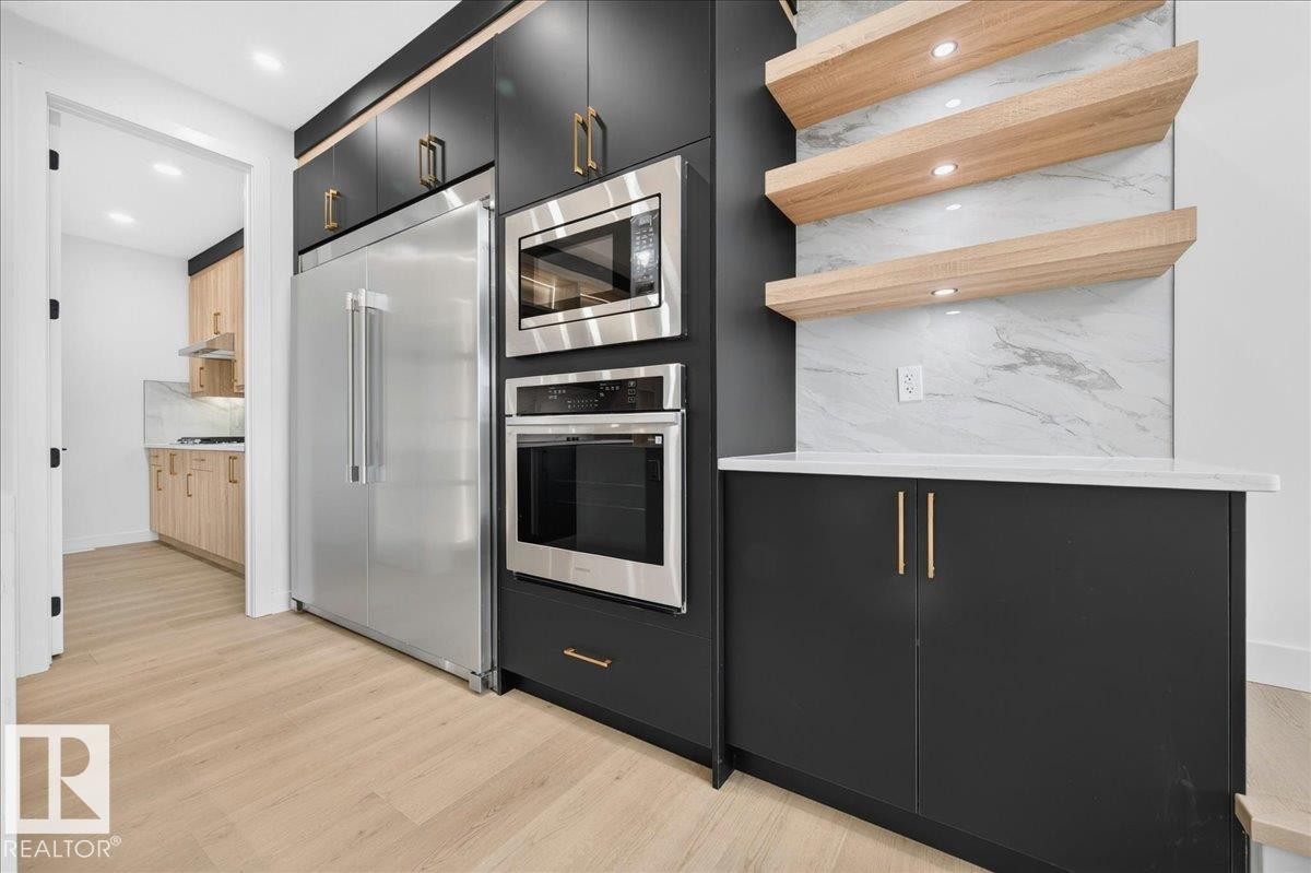 Sleek kitchen featuring dark cabinetry with gold-toned hardware, integrated stainless steel appliances, light wood-finish flooring, and floating shelves above a stone-look backsplash - 403 172 Avenue Nw, Edmonton, AB - Indoor