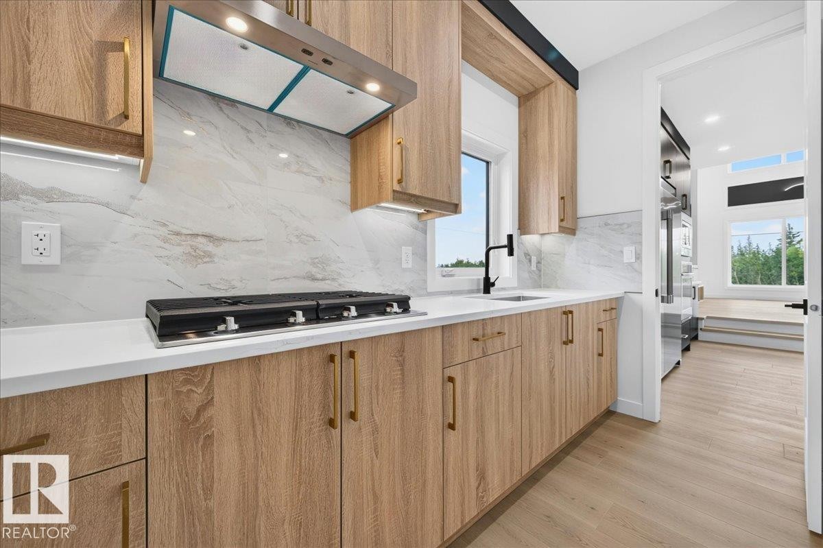 Modern kitchen featuring light wood-finish cabinetry with brass-toned hardware, white solid surface countertops, and a full-height stone-look backsplash - 403 172 Avenue Nw, Edmonton, AB - Indoor Photo Showing Kitchen With Upgraded Kitchen
