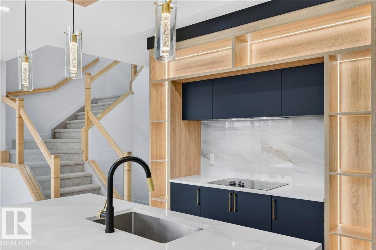 Contemporary kitchen featuring dark blue cabinetry, light wood-finish shelving with integrated lighting, a sleek cooktop, and a white countertop with a stainless steel sink - 403 172 Avenue Nw, Edmonton, AB - Indoor Photo Showing Kitchen With Upgraded Kitchen