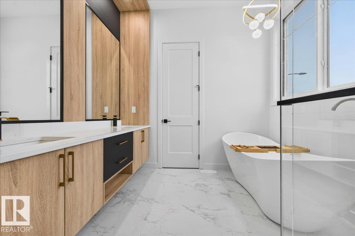 Bathroom featuring a freestanding soaking tub, dual vanity with wood-finish cabinetry, black drawer fronts, gold-tone hardware, and white countertops, large windows, and a modern light fixture - 403 172 Avenue Nw, Edmonton, AB - Indoor Photo Showing Bathroom