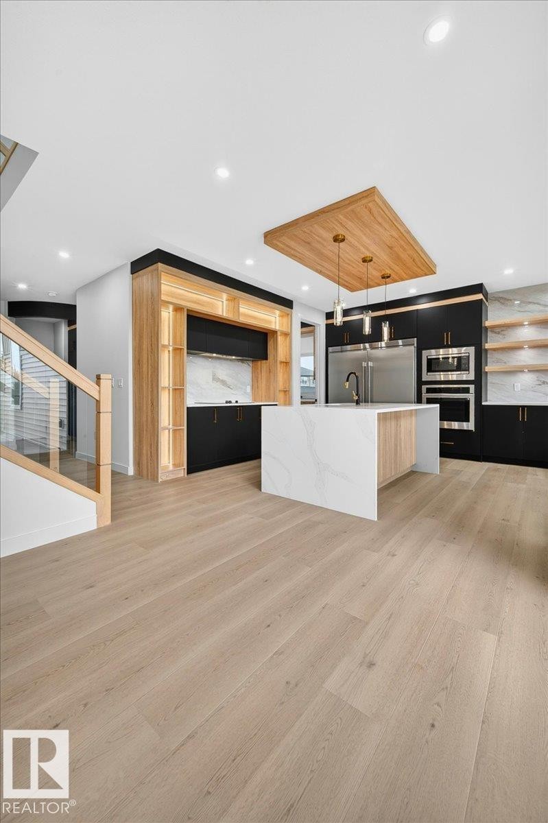 Modern kitchen featuring light wood-finish flooring, a waterfall island with a stone-look countertop, integrated stainless steel appliances, and contrasting black and wood-tone cabinetry - 403 172 Avenue Nw, Edmonton, AB - Indoor