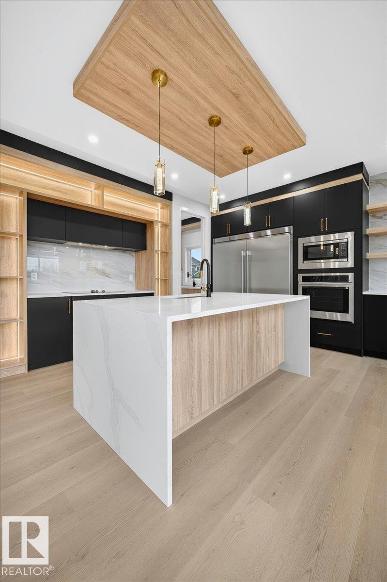 Modern kitchen featuring a waterfall island with a light wood-finish base, sleek black cabinetry, white marble-look backsplash, stainless steel appliances, and light wood-finish flooring - 403 172 Avenue Nw, Edmonton, AB - Indoor Photo Showing Kitchen