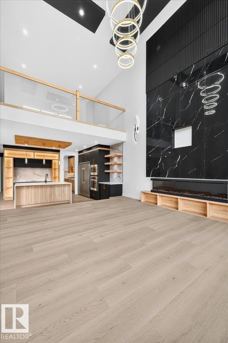 Open-concept living space featuring light wood-finish flooring, a two-story black marble-look accent wall with a linear fireplace, and a kitchen with light wood cabinetry and a central island - 403 172 Avenue Nw, Edmonton, AB - Indoor Photo Showing Other Room