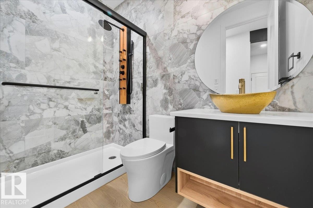 Contemporary bathroom featuring marble-look wall tiling, a gold-toned vessel sink, a dark vanity with gold hardware, a glass-enclosed shower with a black frame, and wood-finish flooring - 403 172 Avenue Nw, Edmonton, AB - Indoor Photo Showing Bathroom