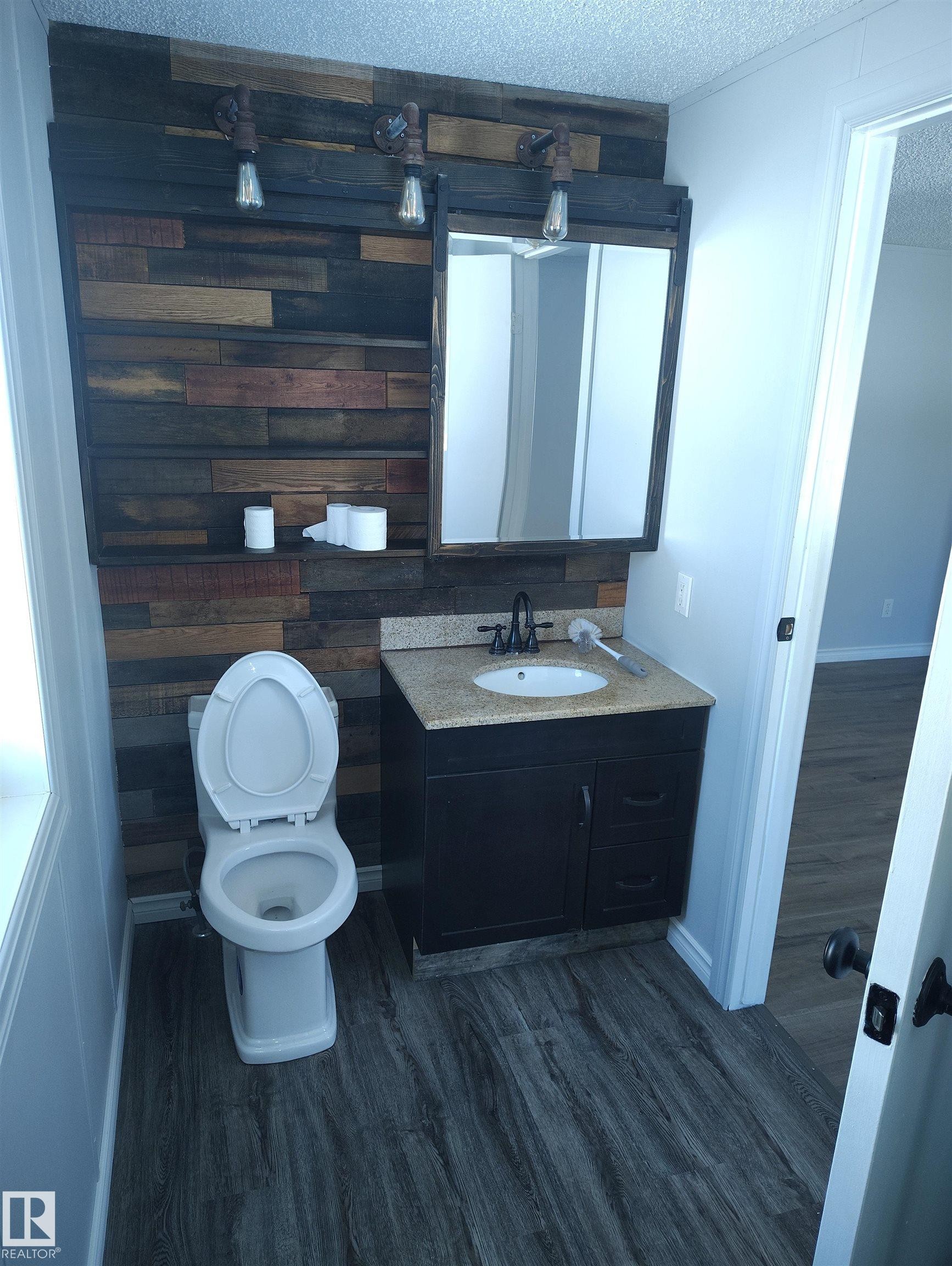 Bathroom featuring a plank wood accent wall with industrial-style sconces, a recessed medicine cabinet, a vanity with a light-colored countertop and integrated sink, and wood-finish flooring - 42 Midland Road, Millet, AB - Indoor Photo Showing Bathroom