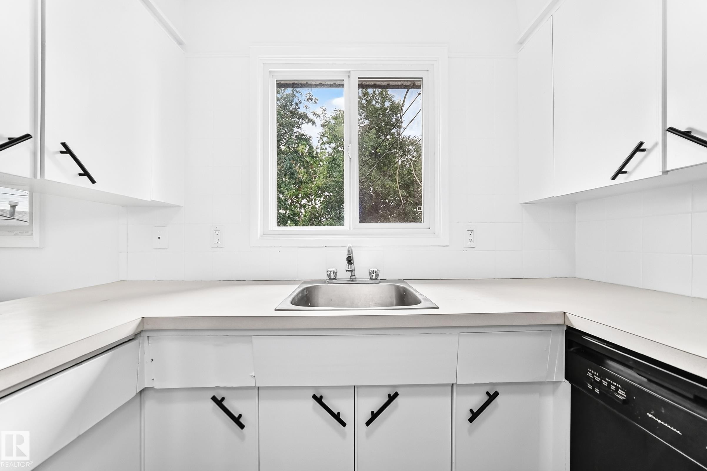 Bright kitchen featuring a stainless steel sink, white cabinetry with matte black hardware, white countertops, and a full-size black dishwasher - 5519 142 Avenue Nw, Edmonton, AB - Indoor Photo Showing Kitchen