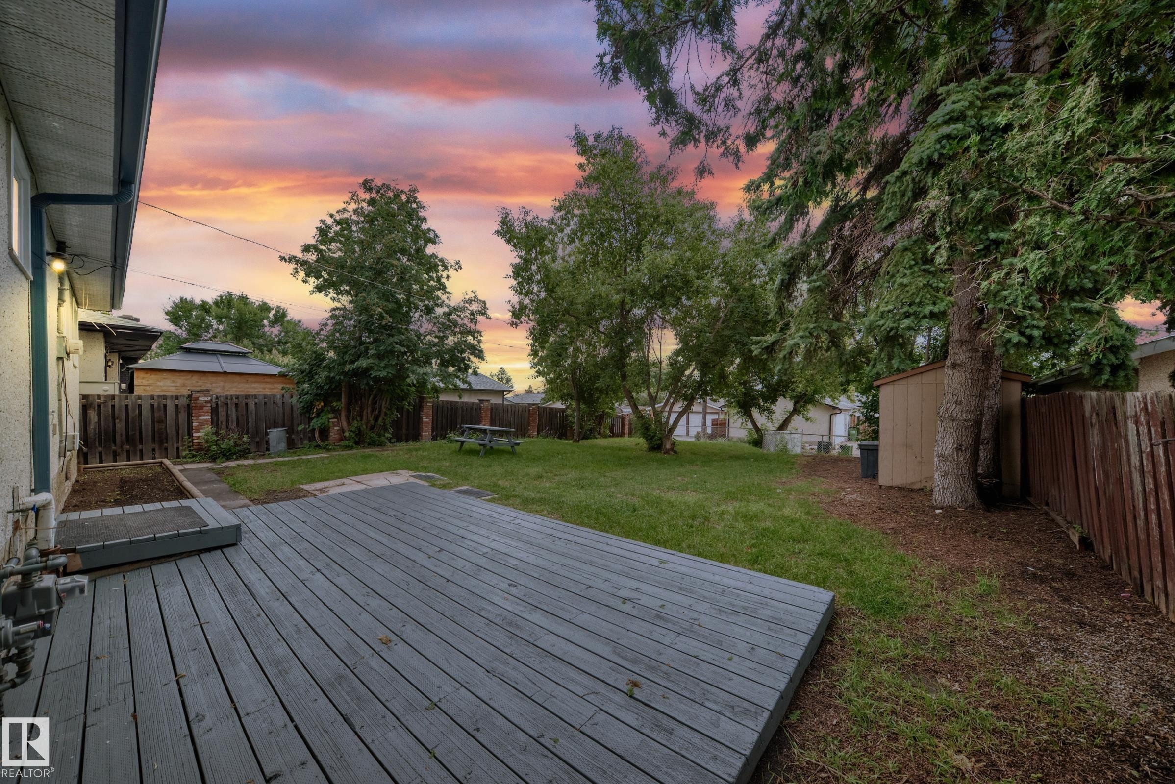 Spacious backyard featuring a large wood-finish deck, expansive lawn, mature perimeter trees, and a wooden storage shed - 5519 142 Avenue Nw, Edmonton, AB - Outdoor With Deck Patio Veranda