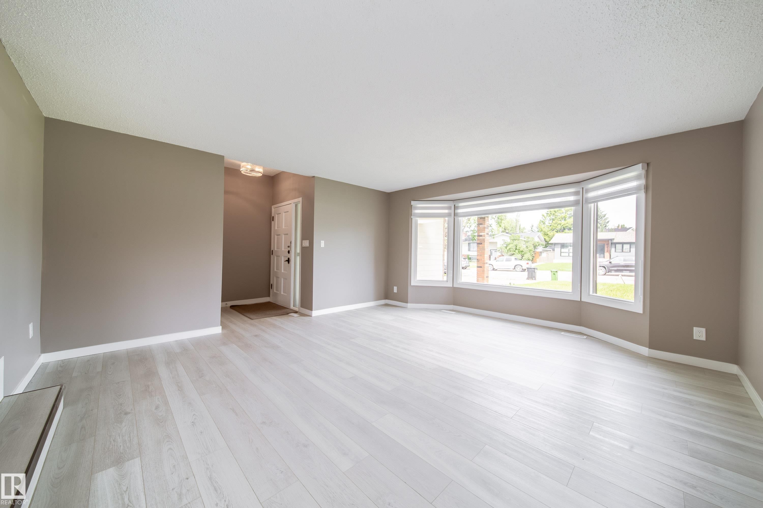 Spacious living area featuring light wood-finish flooring, a large bay window with horizontal blinds, and neutral-toned wall paint - 6907 19 Avenue Nw, Edmonton, AB - Indoor