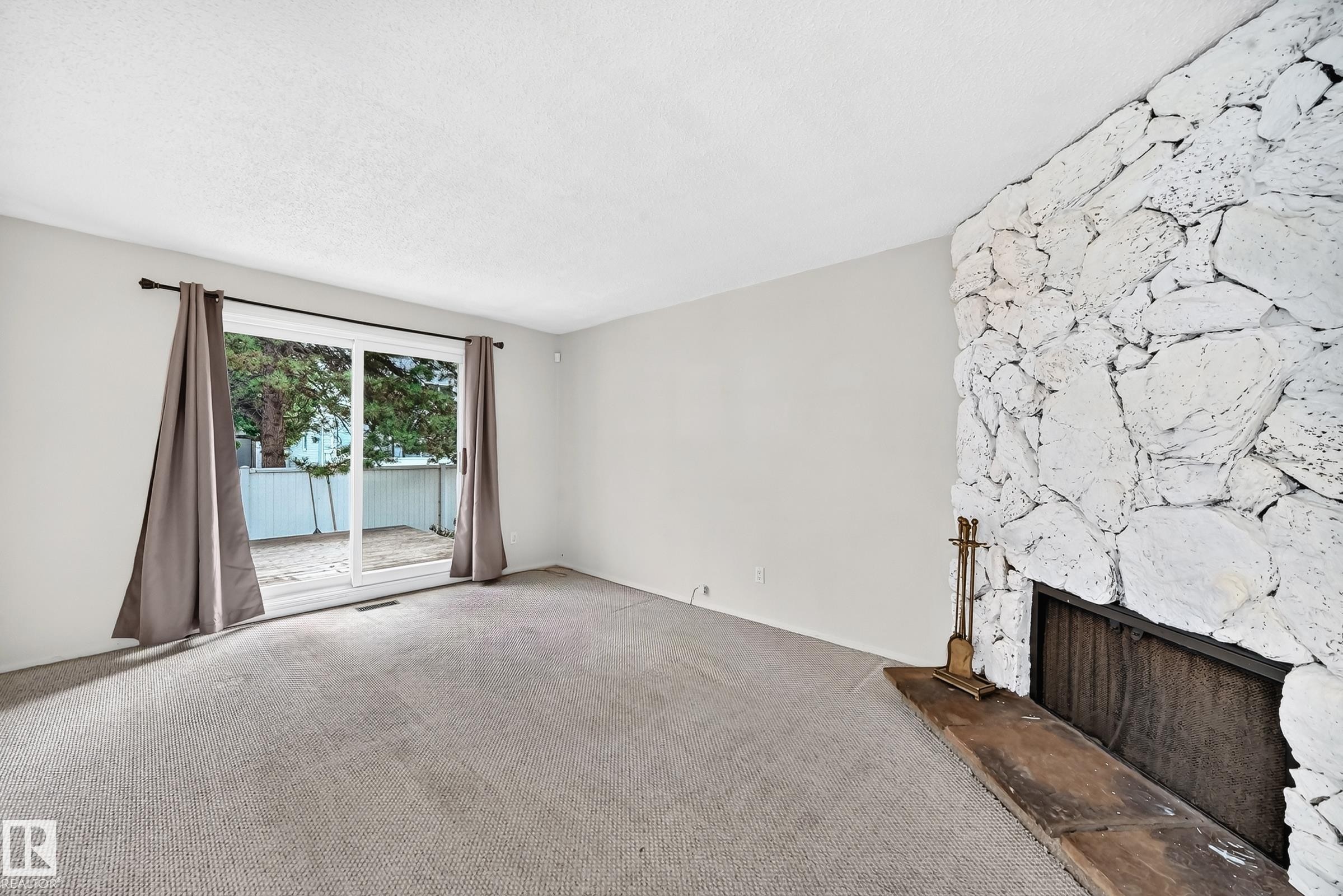 Spacious living room with neutral carpeting, large sliding glass doors opening to outdoor space, and a distinctive white textured stone fireplace feature wall with dark hearth detail - 290 Lancaster Terrace Nw, Edmonton, AB - Indoor With Fireplace