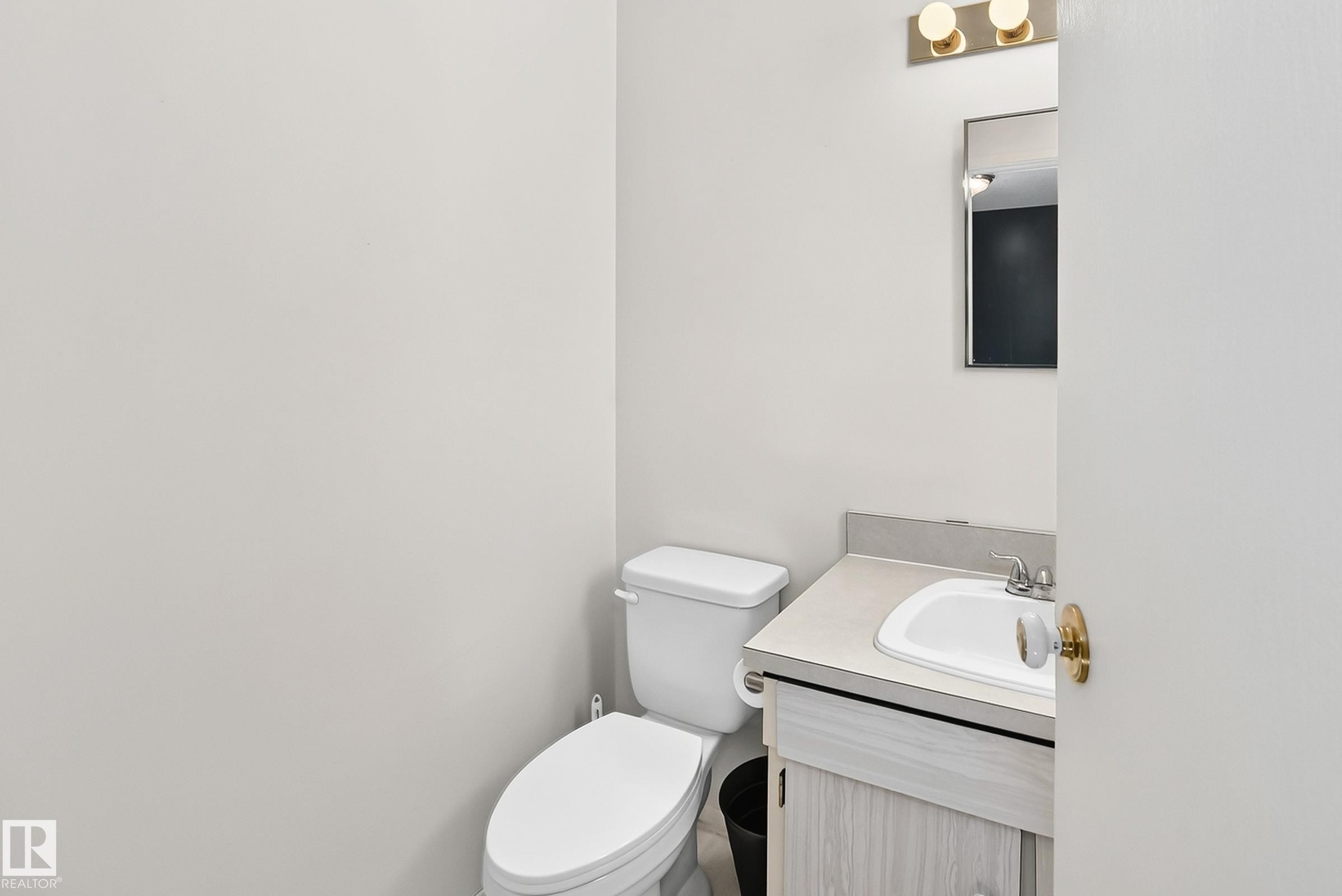 Bathroom featuring a white toilet, light-toned wood-finish vanity, integrated sink, and a wall-mounted mirror with brass-finish lighting - 290 Lancaster Terrace Nw, Edmonton, AB - Indoor Photo Showing Bathroom