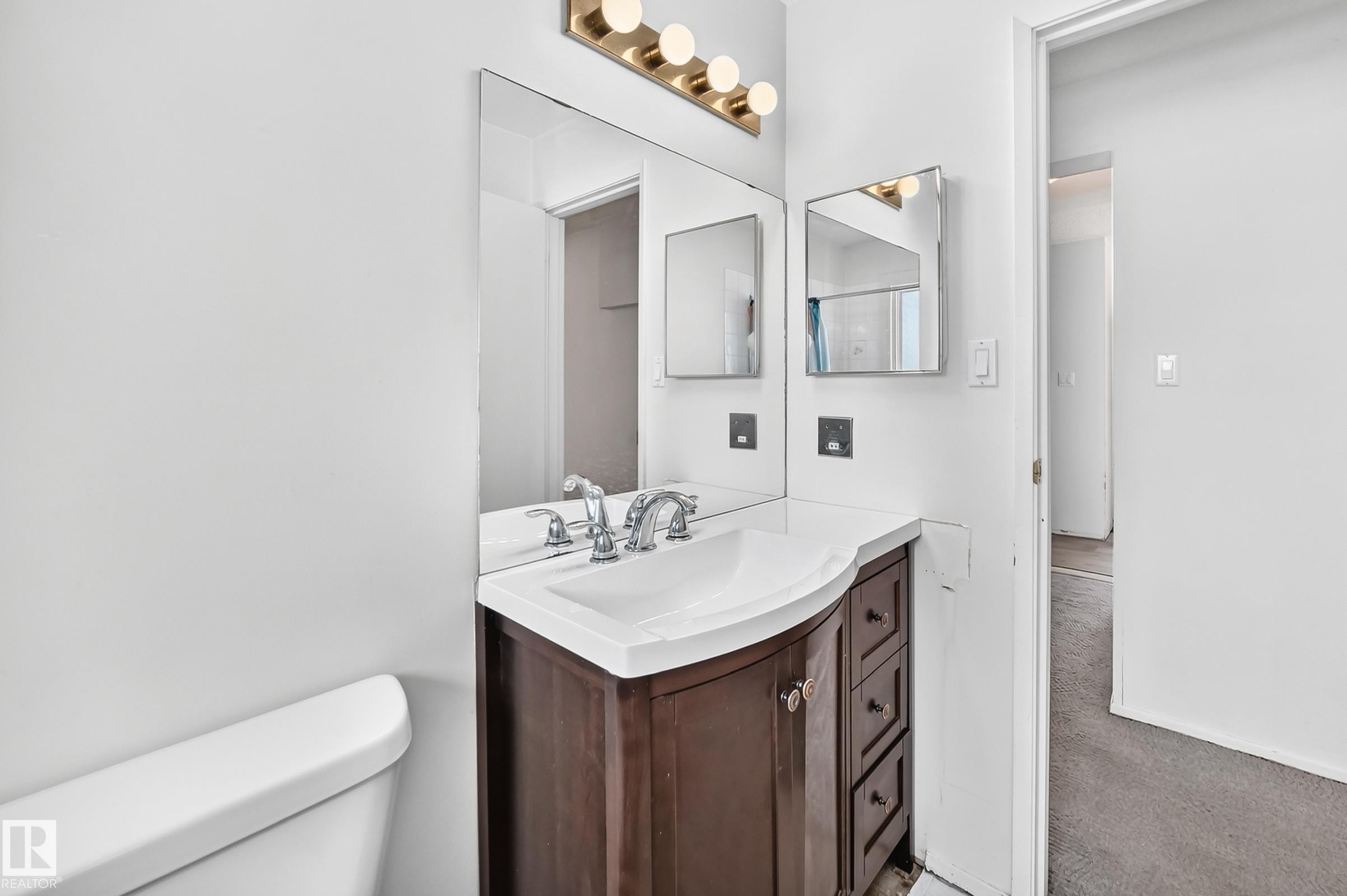 Elegant bathroom vanity featuring a dark wood-finish cabinet with multiple drawers, white porcelain sink, polished chrome faucet fixtures, and ample mirror with brass-finish lighting - 290 Lancaster Terrace Nw, Edmonton, AB - Indoor Photo Showing Bathroom