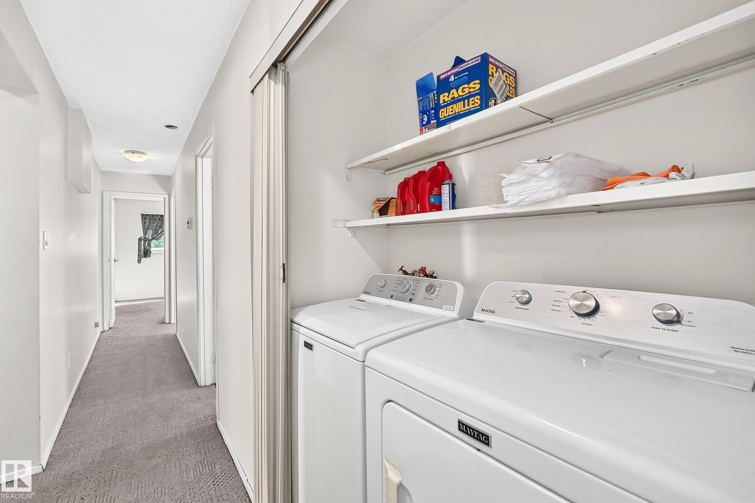 Dedicated laundry room featuring side-by-side washer and dryer with white enamel finish, open shelving with chrome hardware for storage, and gray carpeted flooring - 290 Lancaster Terrace Nw, Edmonton, AB - Indoor Photo Showing Laundry Room