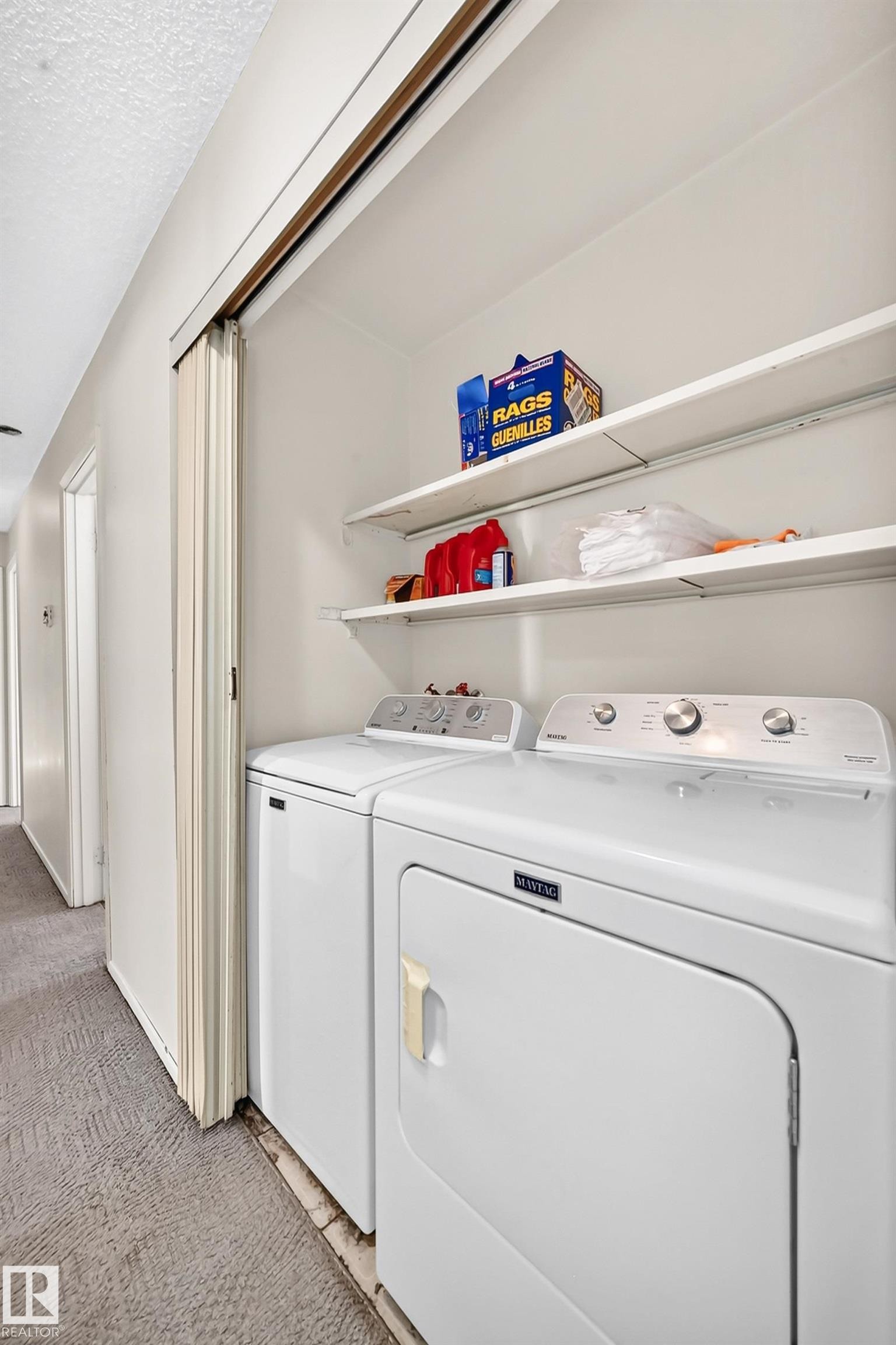Dedicated laundry room featuring side-by-side washer and dryer with stainless steel finishes, open shelving for storage and supplies, and neutral carpeted flooring - 290 Lancaster Terrace Nw, Edmonton, AB - Indoor Photo Showing Laundry Room