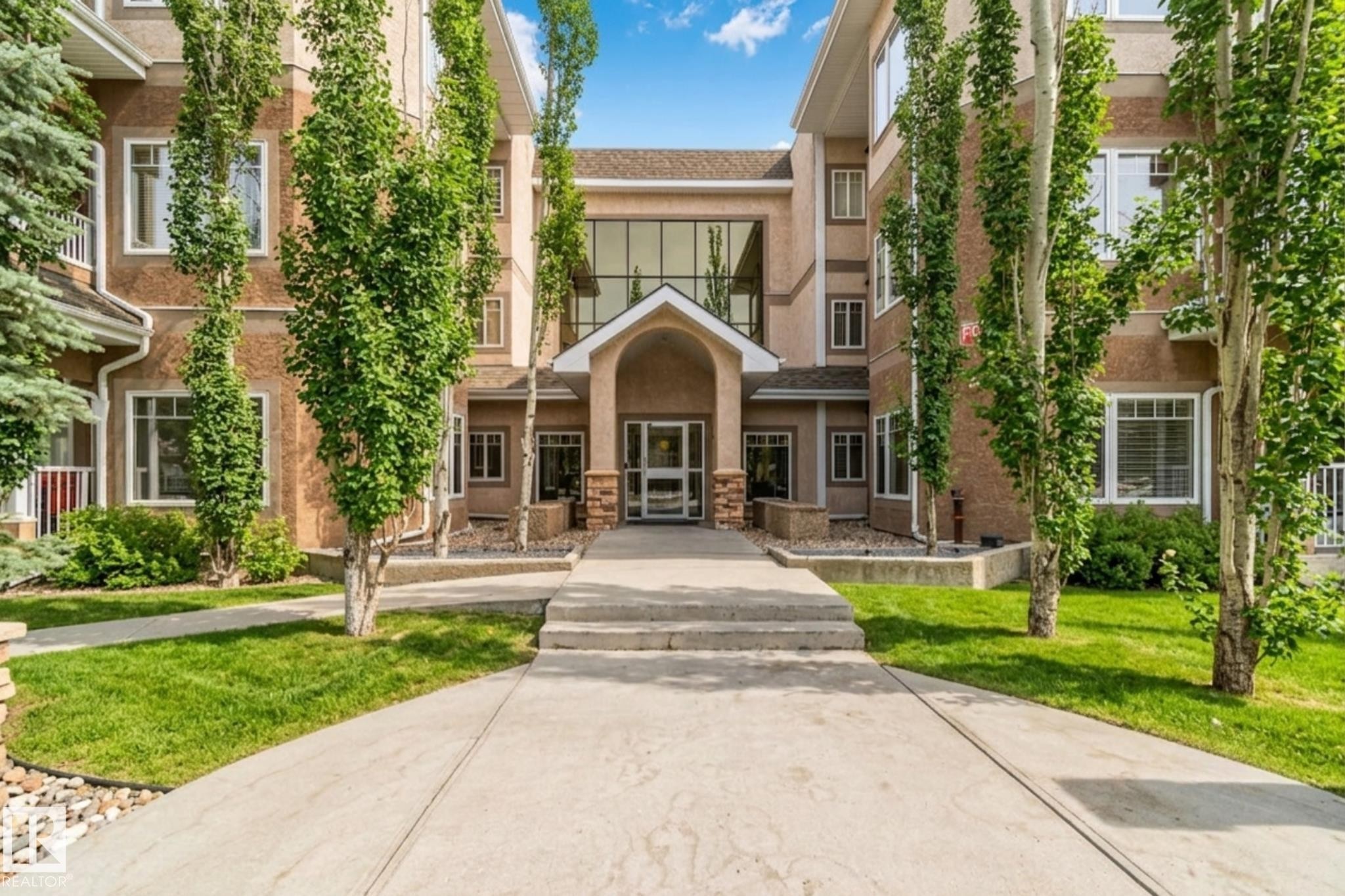Building exterior featuring a stucco facade, multiple windows, a prominent arched entrance, and established landscaping with trees and grass - #120 8931 156 St Nw, Edmonton, AB