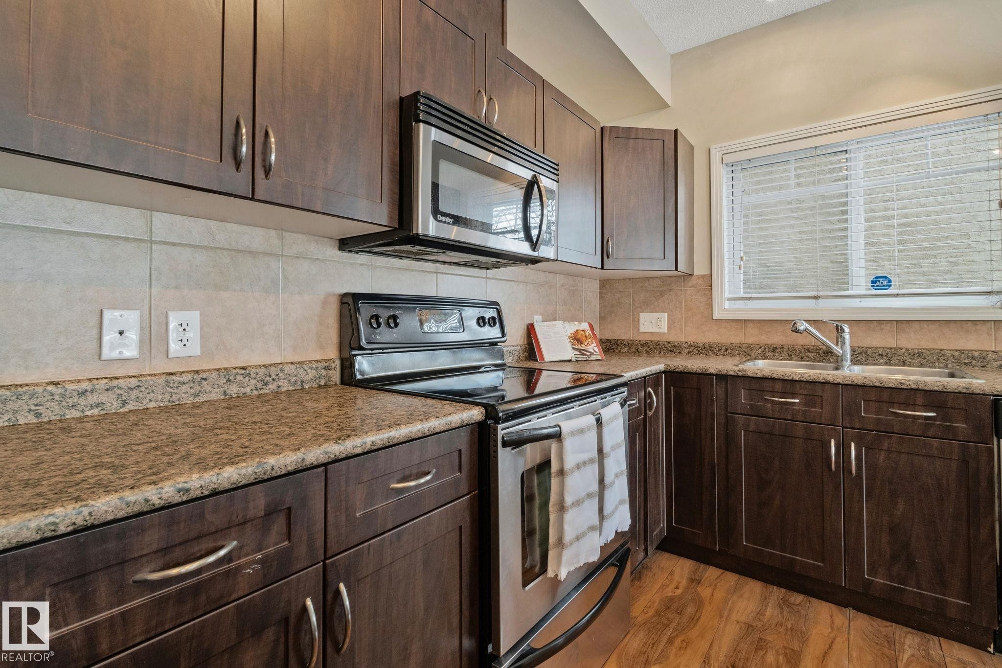 Elegant kitchen featuring rich wood-finish cabinetry with brushed metal hardware, granite countertops with decorative edge banding, and stainless steel appliances including a gas range and over-the-range microwave - #120 8931 156 St Nw, Edmonton, AB