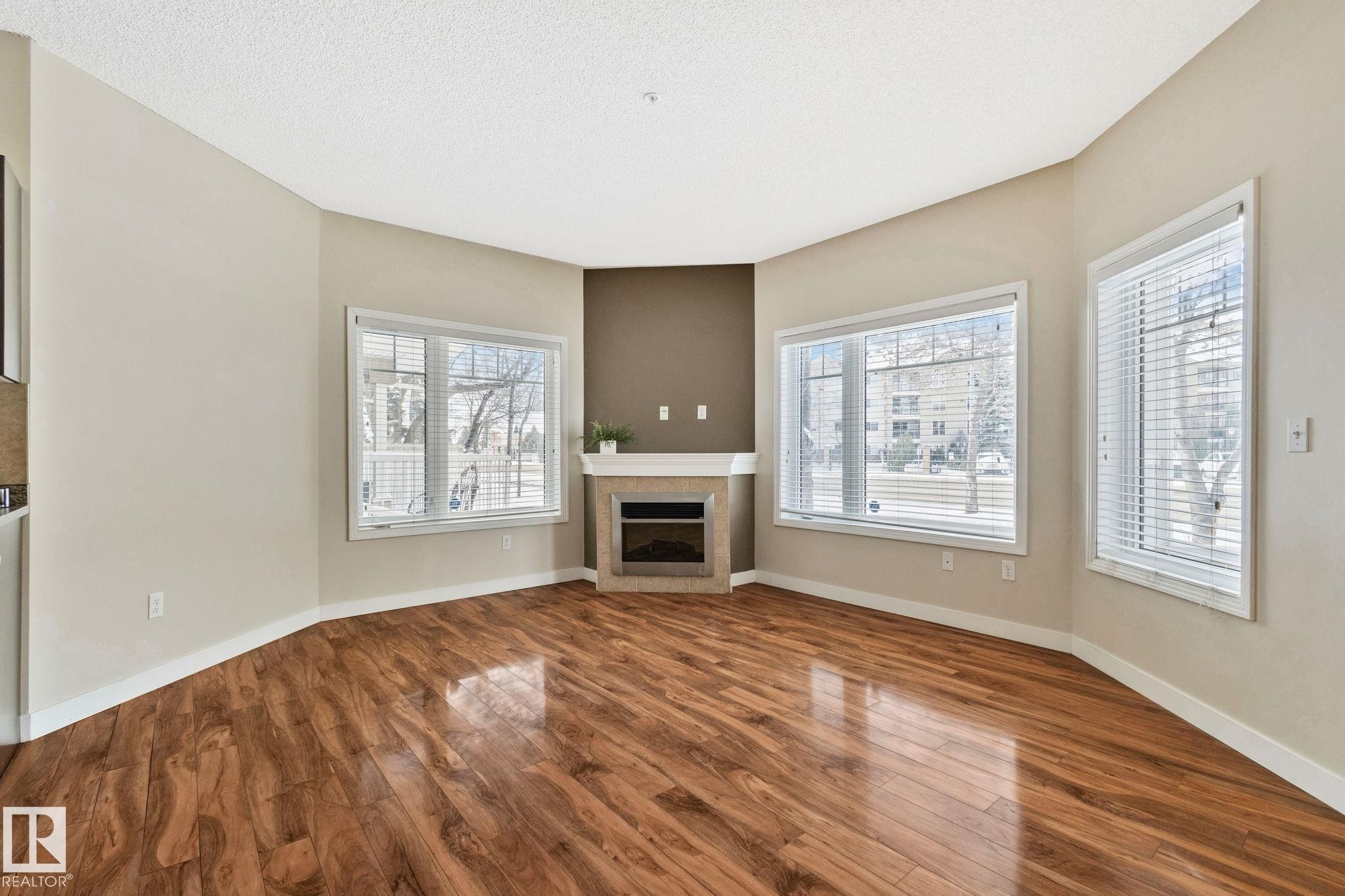 Open living area featuring wood-finish flooring, a corner fireplace with a mantel, and multiple large windows with blinds - #120 8931 156 St Nw, Edmonton, AB