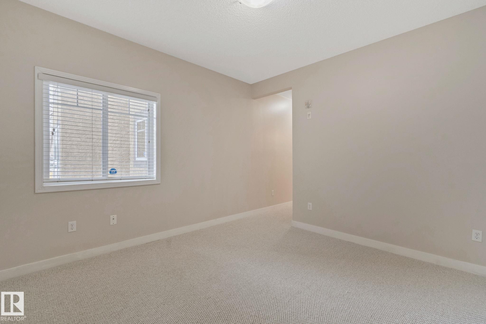Bright secondary bedroom featuring neutral wall finishes, neutral carpet flooring, and abundant natural light from a large window with plantation shutters - #120 8931 156 St Nw, Edmonton, AB