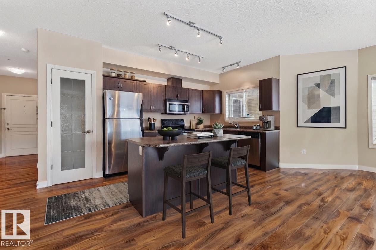 Kitchen featuring wood-finish flooring, dark wood cabinetry, stainless steel appliances, and a central island with bar seating - #120 8931 156 St Nw, Edmonton, AB