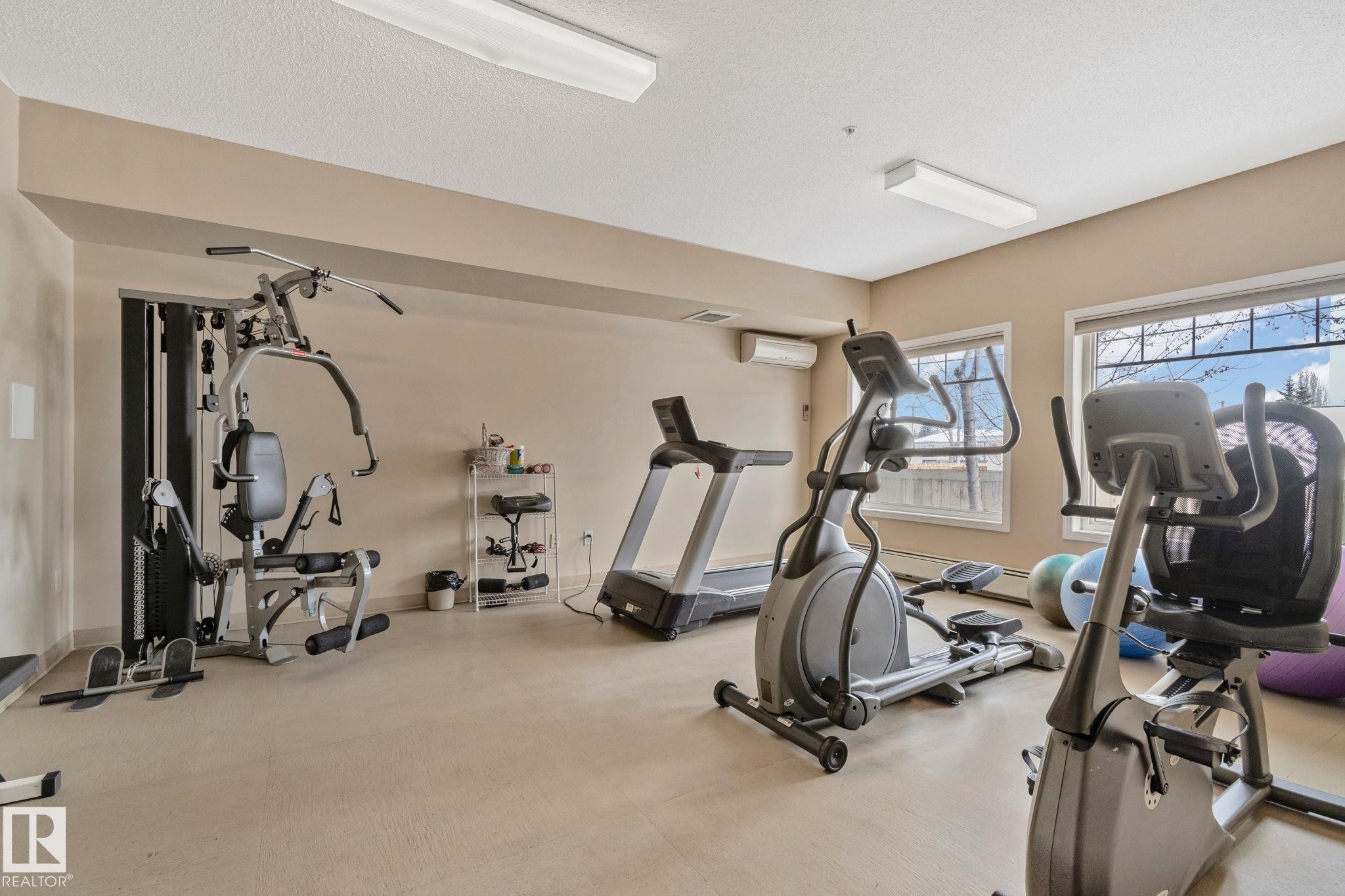 Fitness room featuring two large windows, neutral-toned walls, and overhead fluorescent lighting - #120 8931 156 St Nw, Edmonton, AB