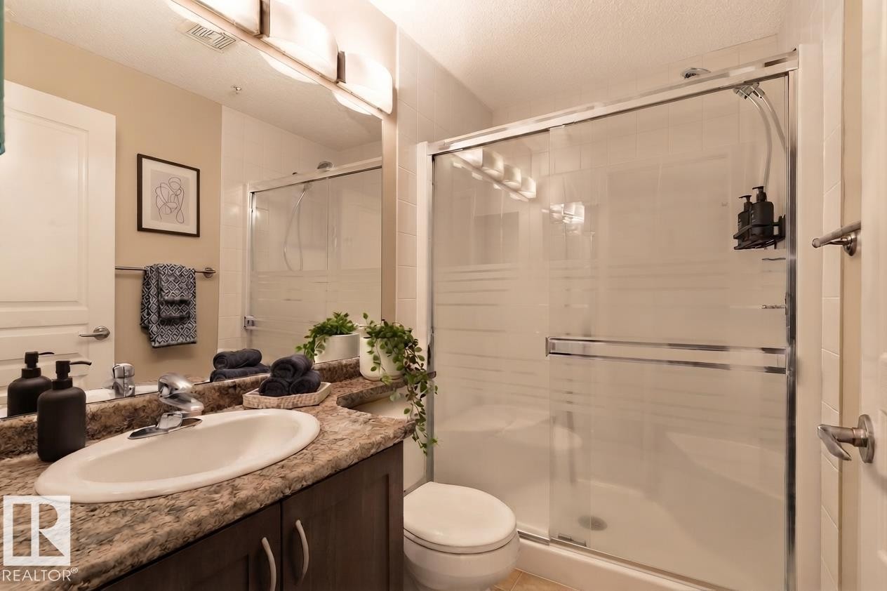Vanity with a patterned countertop and undermount sink, built-in cabinetry, large wall mirror, and a glass-enclosed shower with sliding doors - #120 8931 156 St Nw, Edmonton, AB