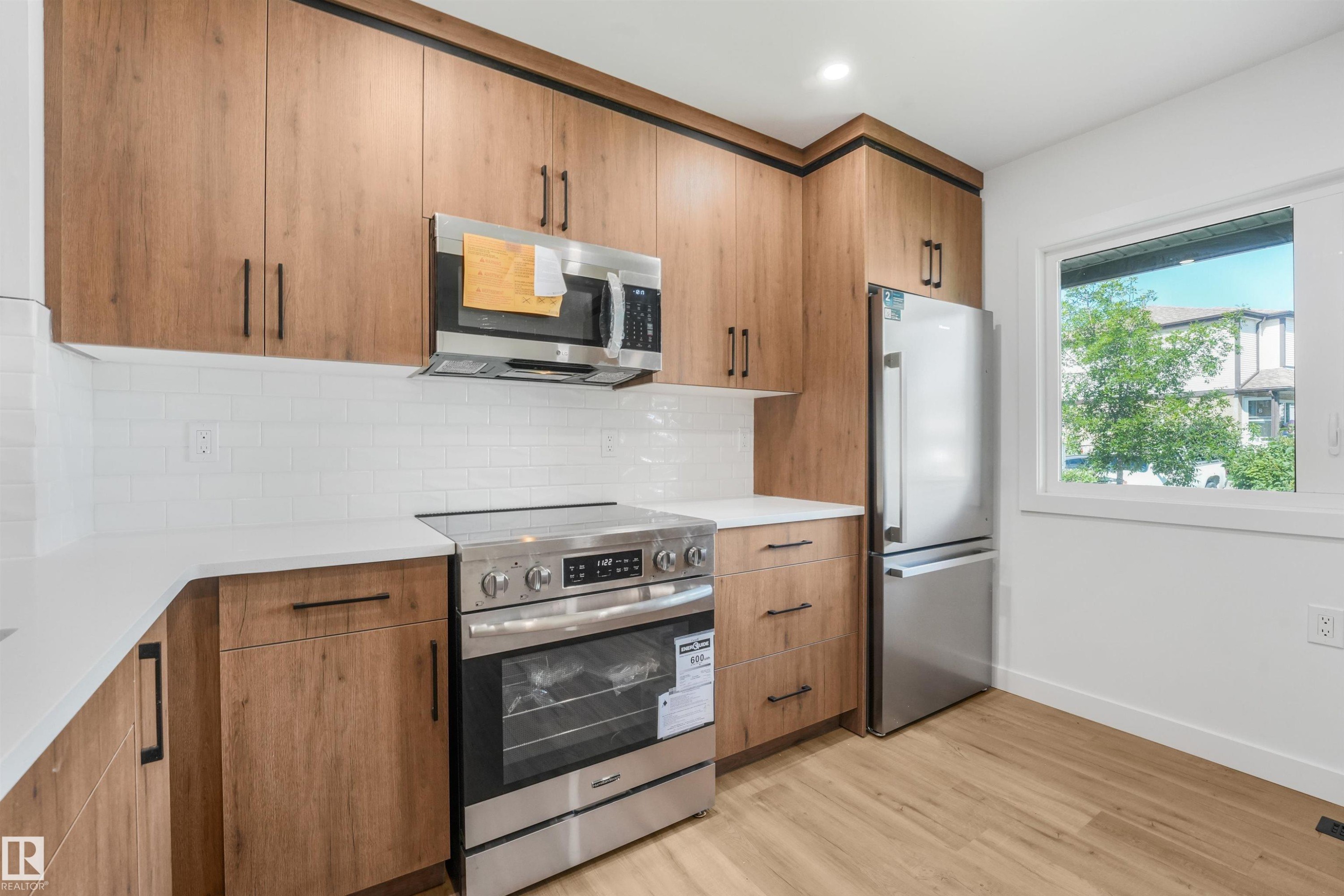 Modern kitchen featuring wood-finish cabinetry with black hardware, stainless steel appliances, white subway tile backsplash, and light countertops - #39 14511 52 Street Nw, Edmonton, AB - Indoor Photo Showing Kitchen