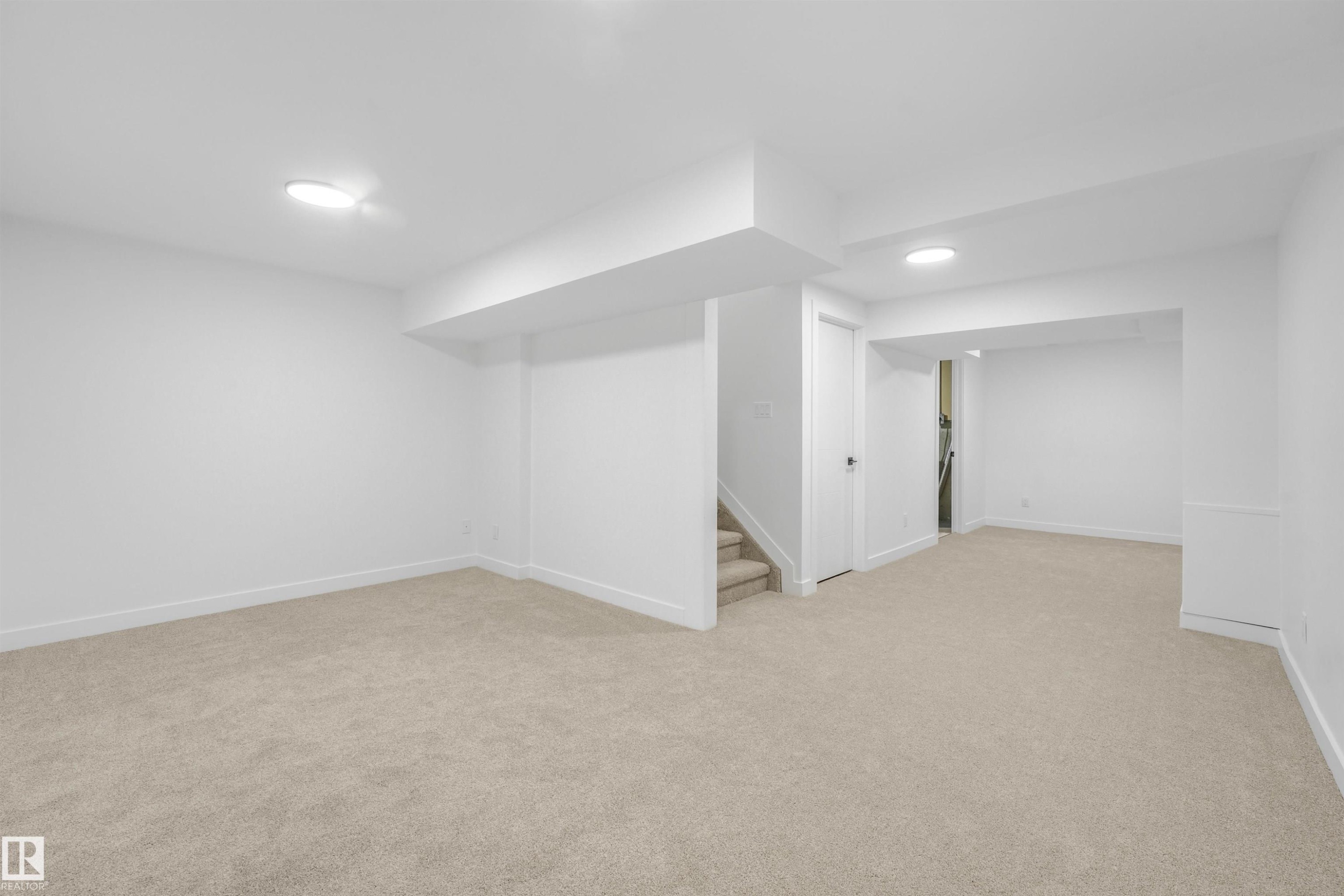 Spacious lower-level room featuring light-toned carpeting, crisp white walls, recessed ceiling lights, a white paneled door, and a carpeted staircase - #39 14511 52 Street Nw, Edmonton, AB - Indoor