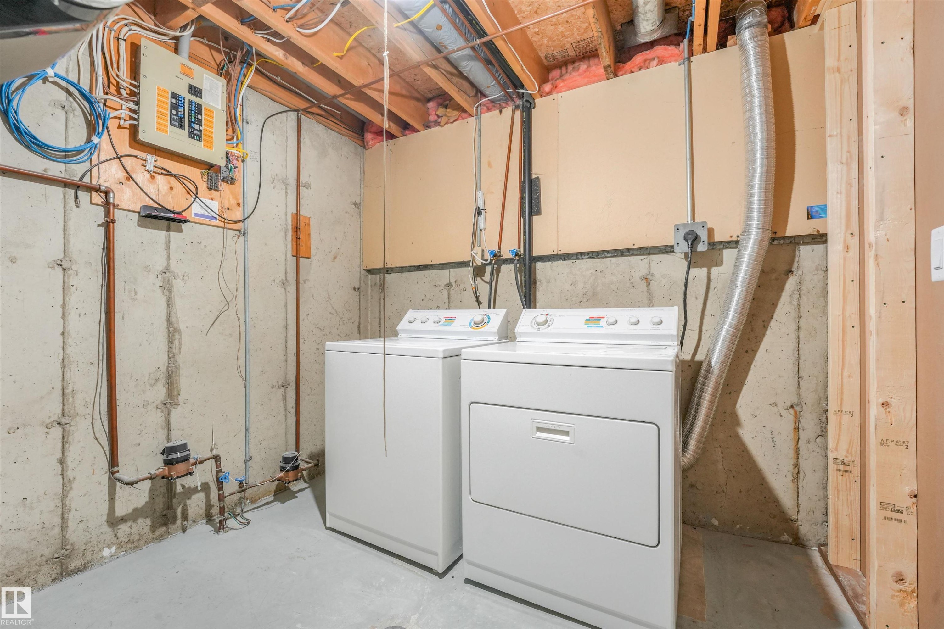 Dedicated laundry area featuring a white washing machine and dryer, exposed concrete walls, and visible utility infrastructure including copper piping and electrical panel - #39 14511 52 Street Nw, Edmonton, AB - Indoor Photo Showing Laundry Room
