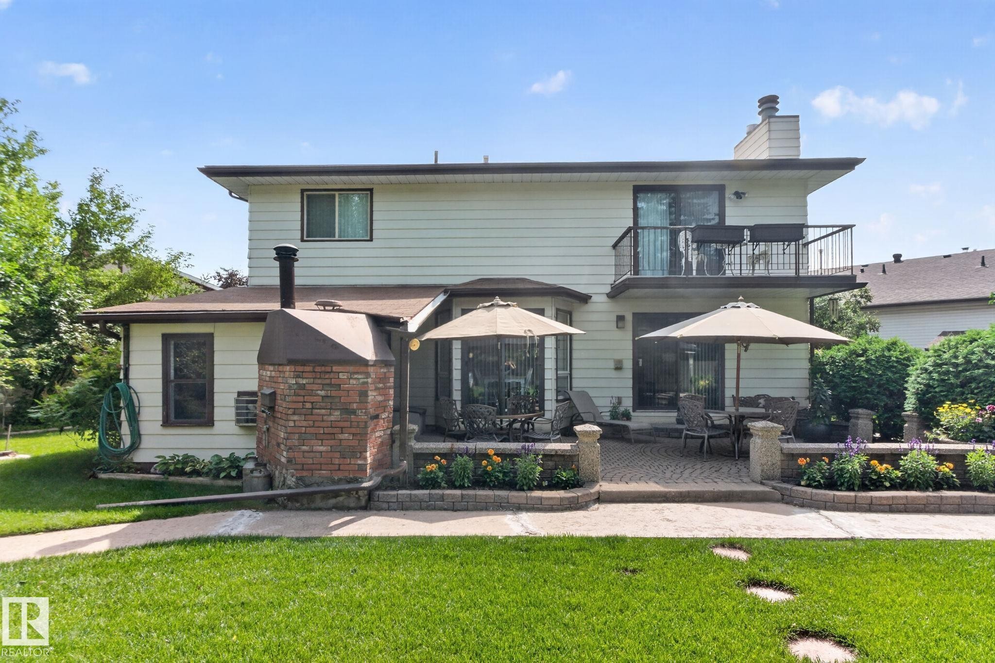 Expansive backyard featuring a manicured lawn, a two-tier paver patio, a built-in brick outdoor fireplace, and raised garden beds with stone caps - 18132 78 Avenue Nw, Edmonton, AB - Outdoor With Balcony With Deck Patio Veranda