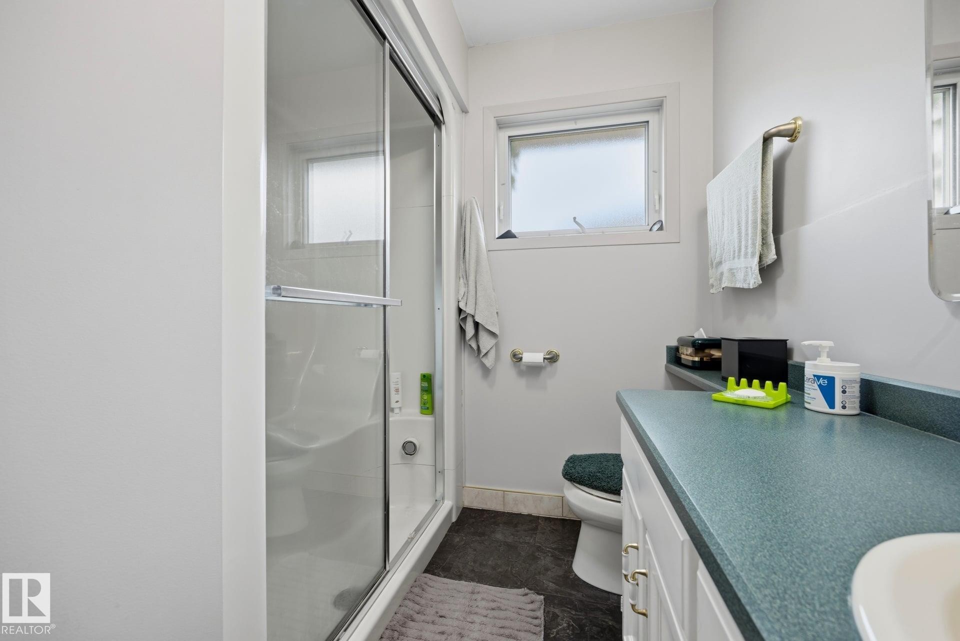 Enclosed shower with glass sliding door, vanity with integrated sink, white cabinetry with brass hardware, and a small window - 9503 54 Street Nw, Edmonton, AB