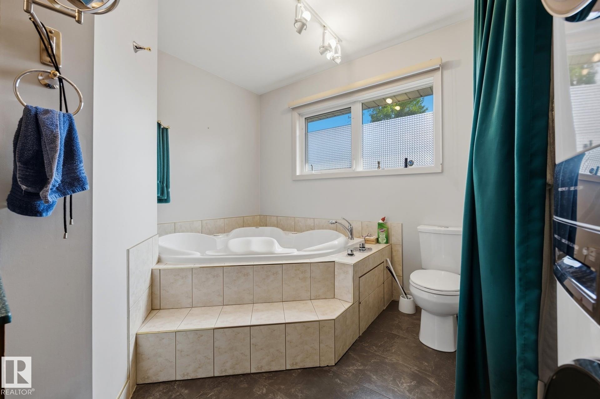 Master bathroom featuring a corner soaking tub with tile surround, separate toilet, pedestal sink with chrome fixtures, tile flooring, and natural light from dual windows with privacy glass - 9503 54 Street Nw, Edmonton, AB