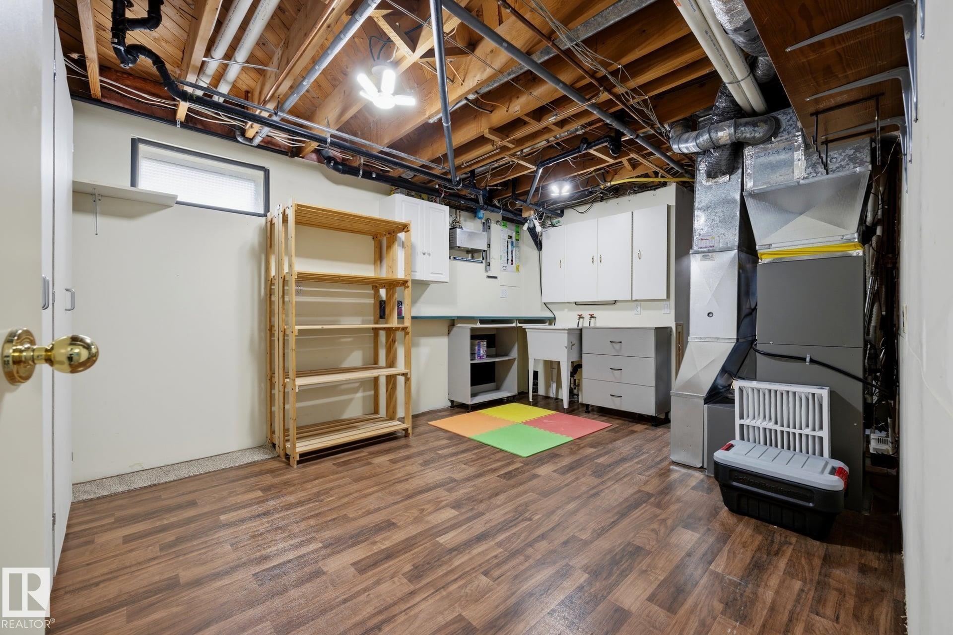 Basement utility area featuring wood-finish flooring, exposed ceiling joists, white cabinetry, a utility sink, and HVAC systems - 9503 54 Street Nw, Edmonton, AB