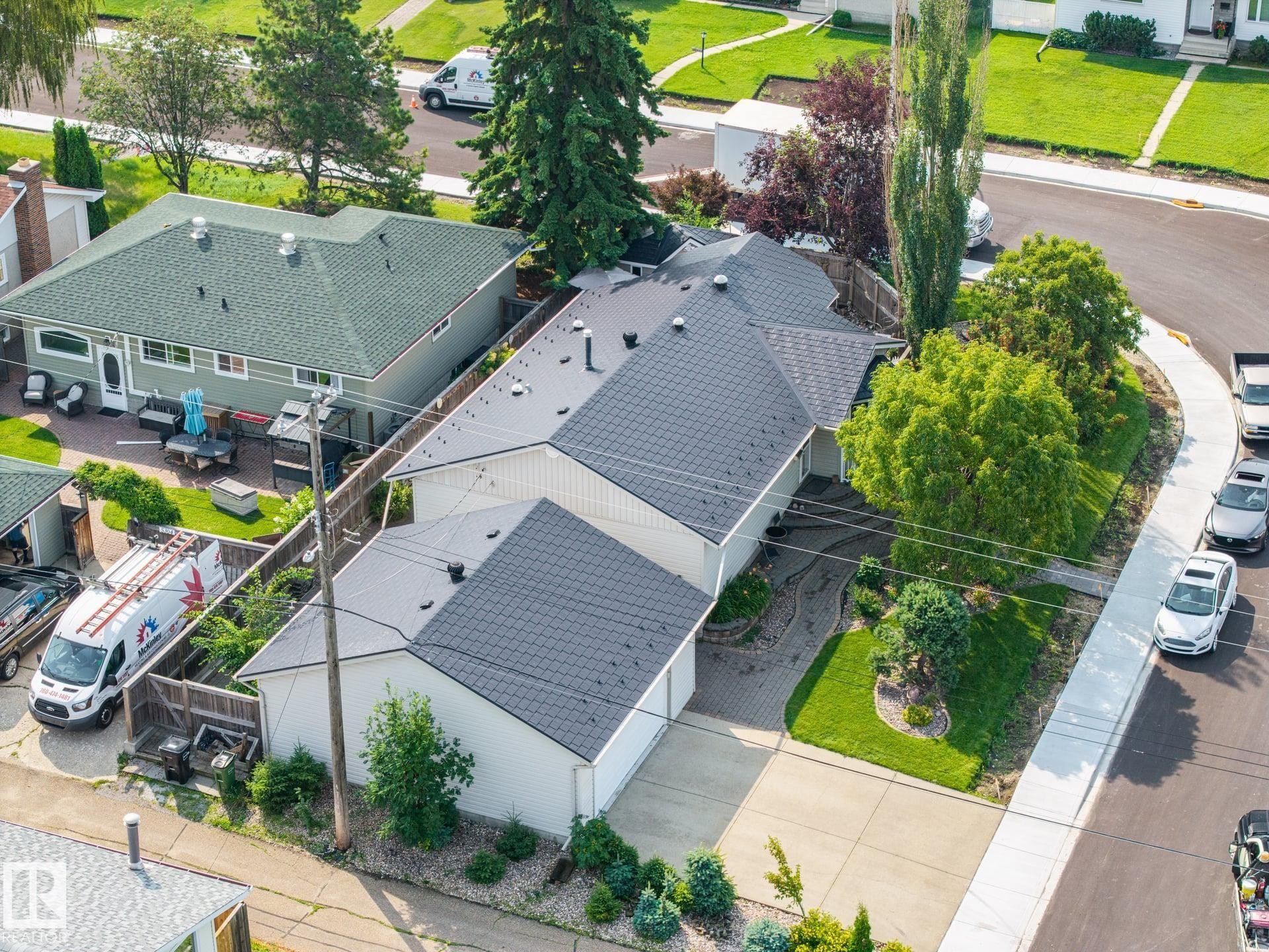 Modern residence featuring a distinctive multi-pitched architectural design with dark asphalt shingle roofing, clean white exterior siding, and professional landscaping with mature greenery throughout the front grounds - 9503 54 Street Nw, Edmonton, AB