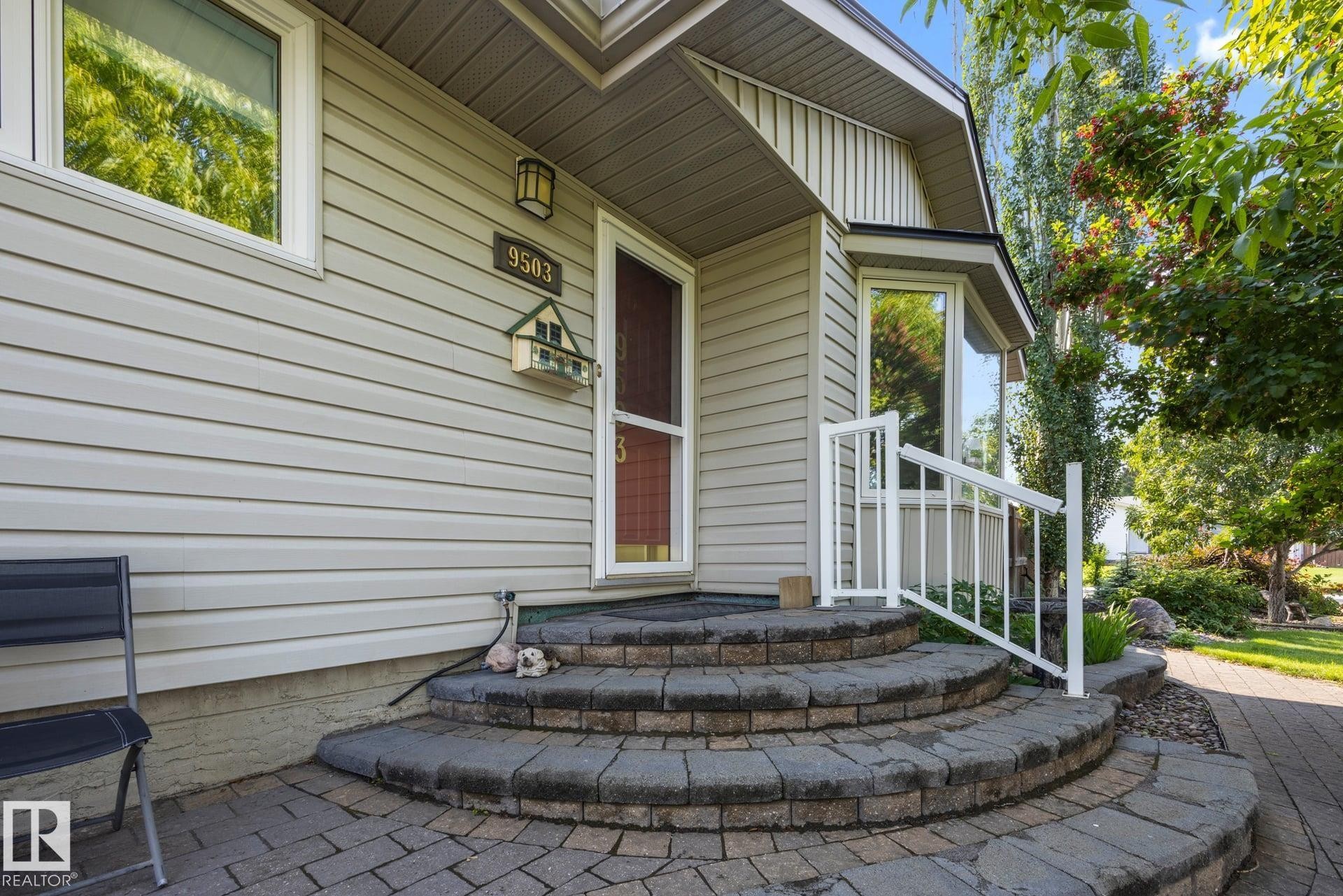 Welcoming front entry featuring a curved paver patio with dimensional stone borders, white railing, and siding exterior accented by landscaped greenery - 9503 54 Street Nw, Edmonton, AB