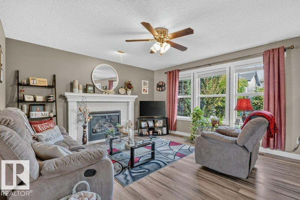 Living area featuring a fireplace with a white mantel, large windows with natural light, wood-finish flooring, and a ceiling fan - 5322 Terwillegar Boulevard Nw, Edmonton, AB - Indoor Photo Showing Living Room With Fireplace