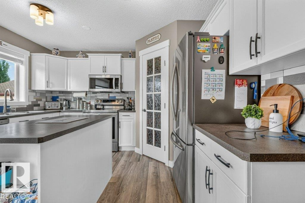 Kitchen featuring white cabinetry, dark countertops, wood-finish flooring, stainless steel appliances, and a glass-paneled pantry door - 5322 Terwillegar Boulevard Nw, Edmonton, AB - Indoor Photo Showing Kitchen With Double Sink