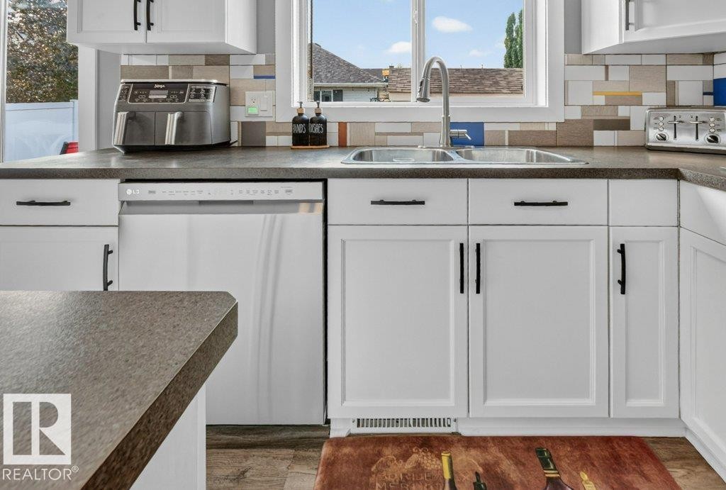 Kitchen featuring white cabinetry with black hardware, dark countertops, an undermount double basin sink with a gooseneck faucet, a built-in dishwasher, and a multi-toned tile backsplash - 5322 Terwillegar Boulevard Nw, Edmonton, AB - Indoor Photo Showing Kitchen With Double Sink