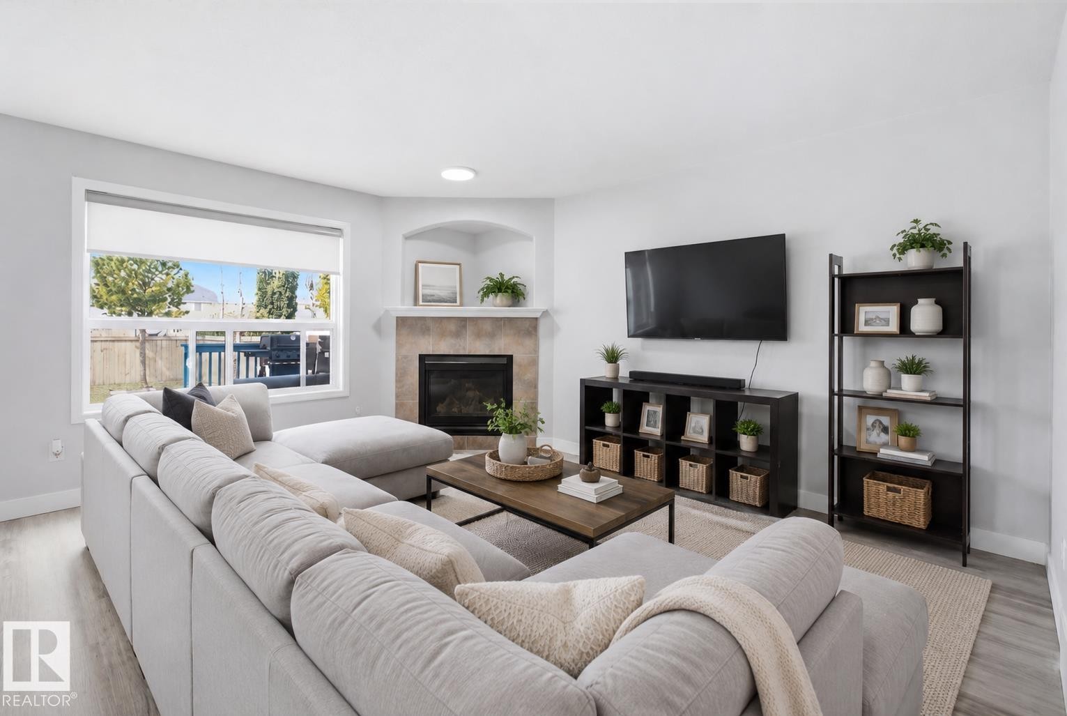 Living room featuring light wood-finish flooring, a corner fireplace with stone tile surround and mantel, a large window with roller shades, and light gray wall paint - 714 87 Street Sw, Edmonton, AB - Indoor Photo Showing Living Room With Fireplace