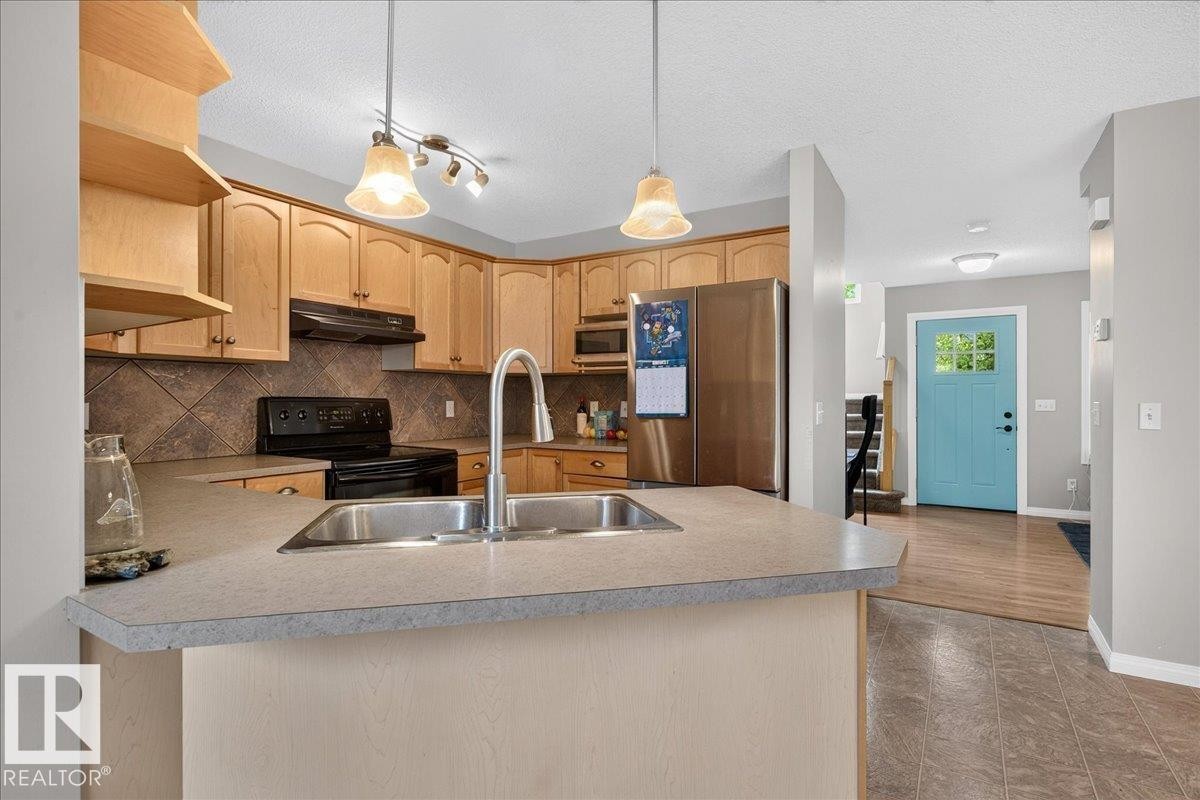 Open-concept kitchen featuring wood-finish cabinetry, stainless steel appliances, and a spacious island with a dual-basin sink and pendant lighting - 887 Oakland Boulevard, Devon, AB - Indoor Photo Showing Kitchen With Double Sink