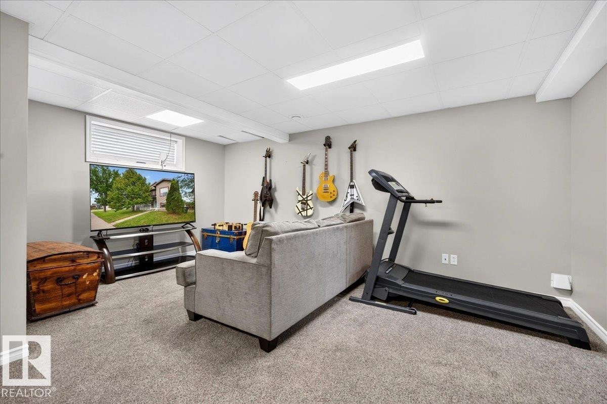 Versatile recreation room featuring neutral carpeting, recessed and linear ceiling lighting, built-in wall-mounted guitar display with multiple hanging mounts, gray upholstered seating, wood-finish media console, wall-mounted television, and a treadmill - 887 Oakland Boulevard, Devon, AB - Indoor Photo Showing Gym Room