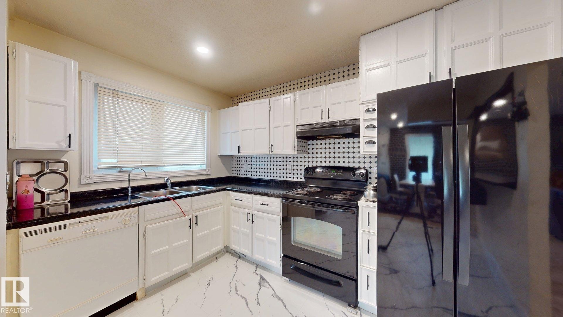 Kitchen featuring white cabinetry, dark countertops, black appliances, a double basin sink, and a patterned backsplash - 12812 45 Street Nw, Edmonton, AB - Indoor Photo Showing Kitchen With Double Sink